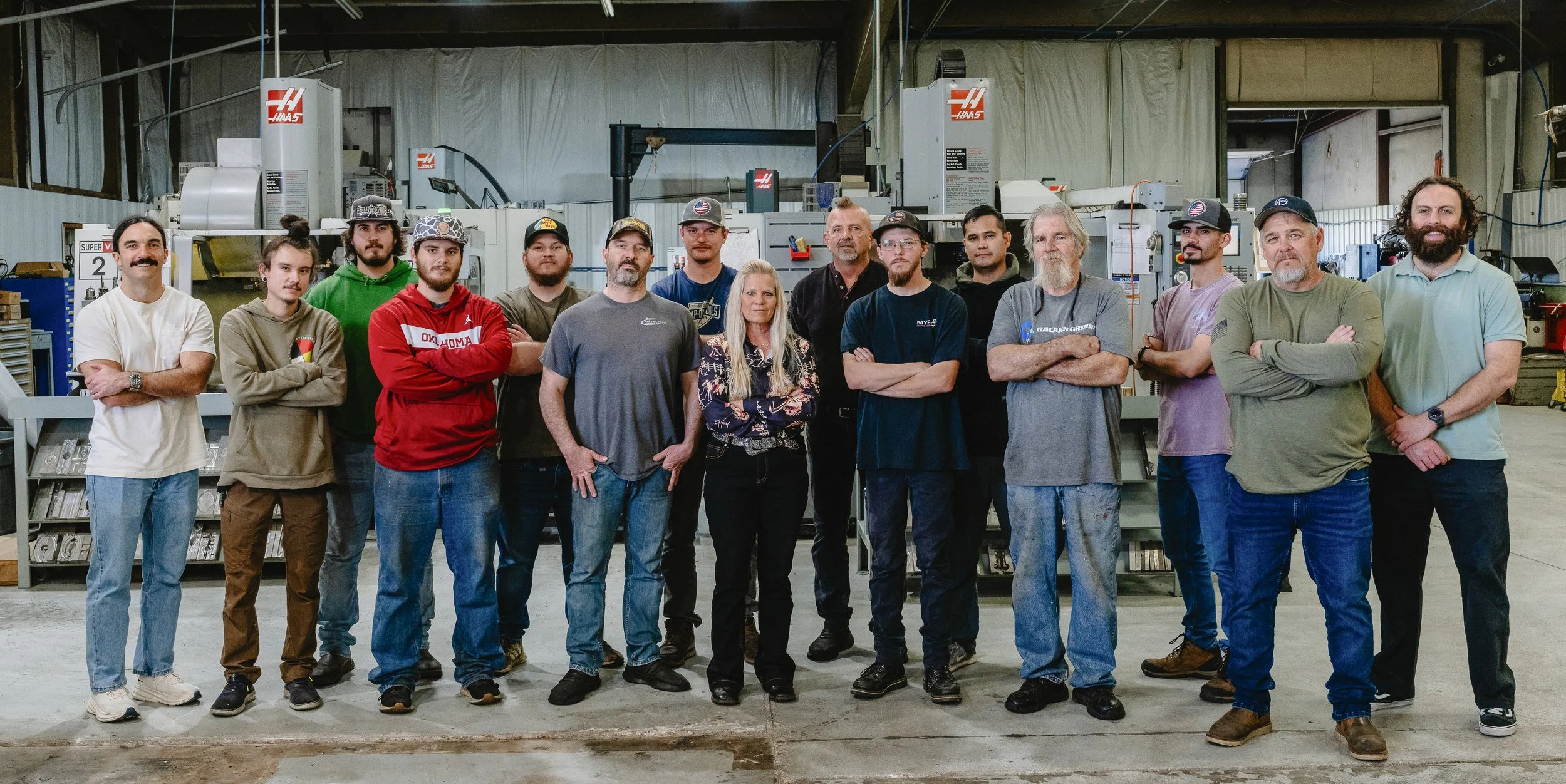 Group of 15 people standing in a factory or industrial setting with machinery in the background.