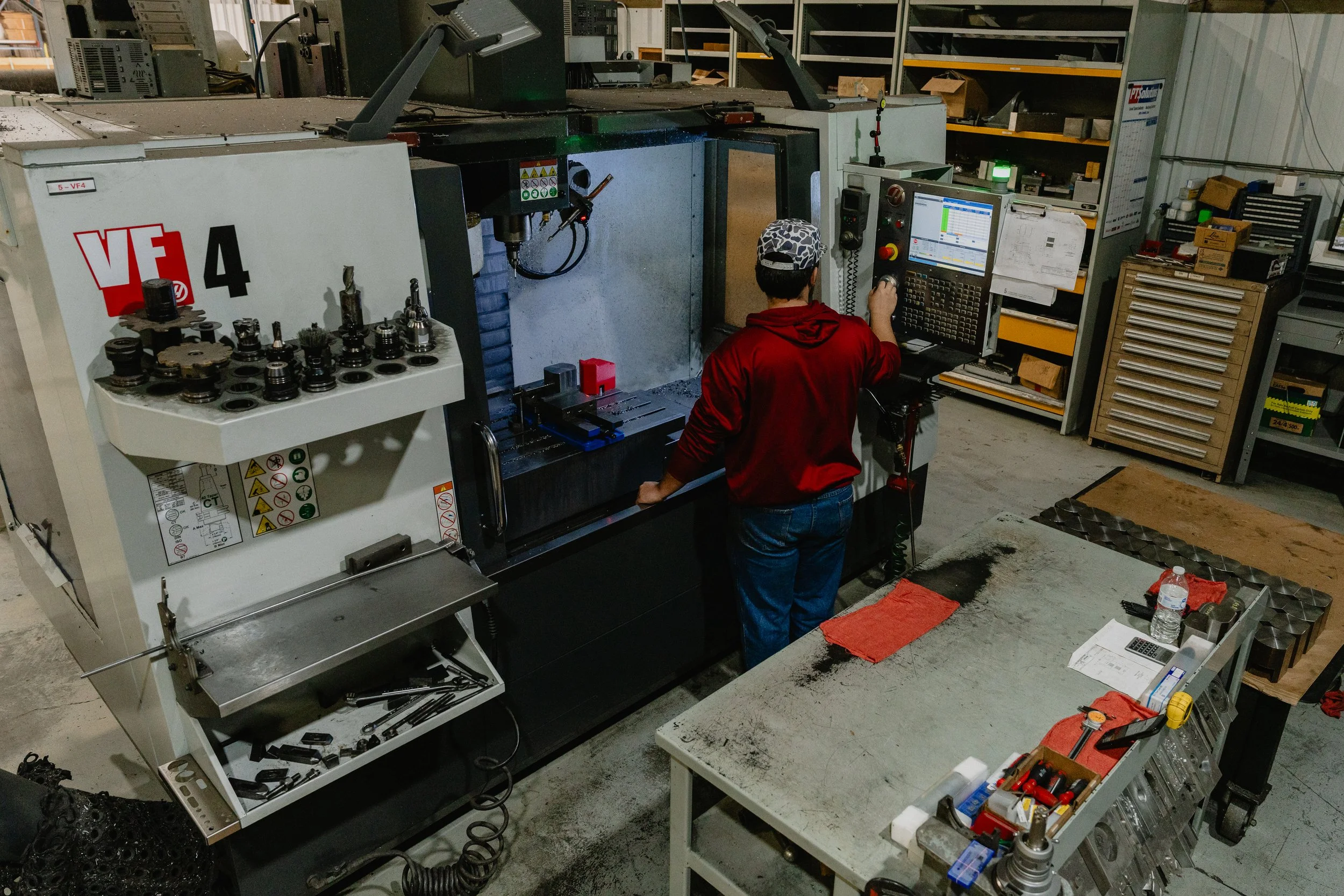 A person wearing a red hoodie and patterned cap operating a CNC machine in a workshop, with tools and equipment on surrounding shelves and workbenches.