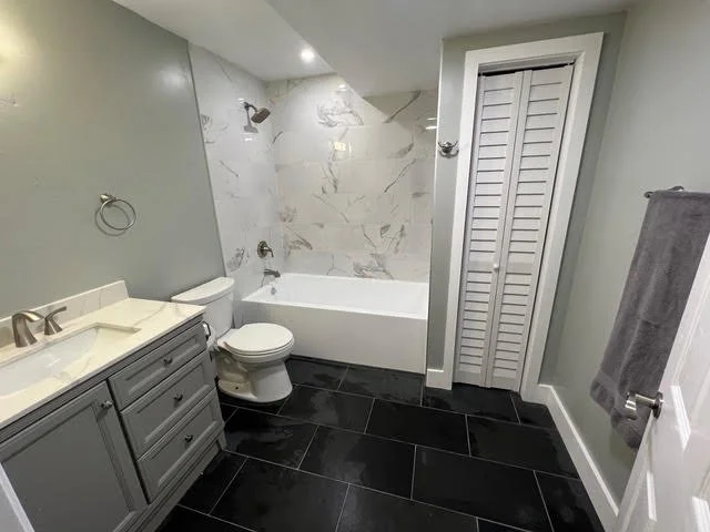 Bathroom with gray vanity, white sink, toilet, bathtub with marble walls, louvered door closet, gray towel on towel rack, black tile floors, and gray wall paint.