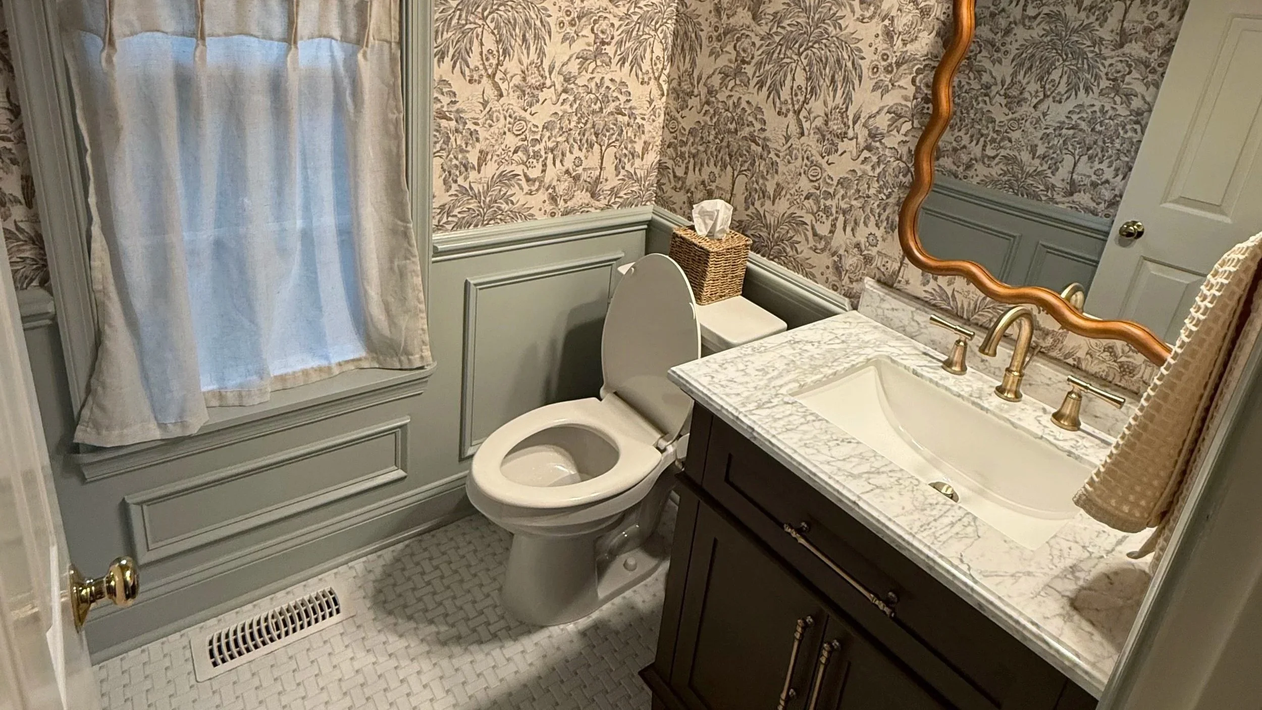 A small bathroom with patterned wallpaper, a window with curtains, a toilet with a wicker tissue box on top, a granite countertop with a sink, a brass faucet, a large ornate mirror, and a wicker towel hanging on the wall.