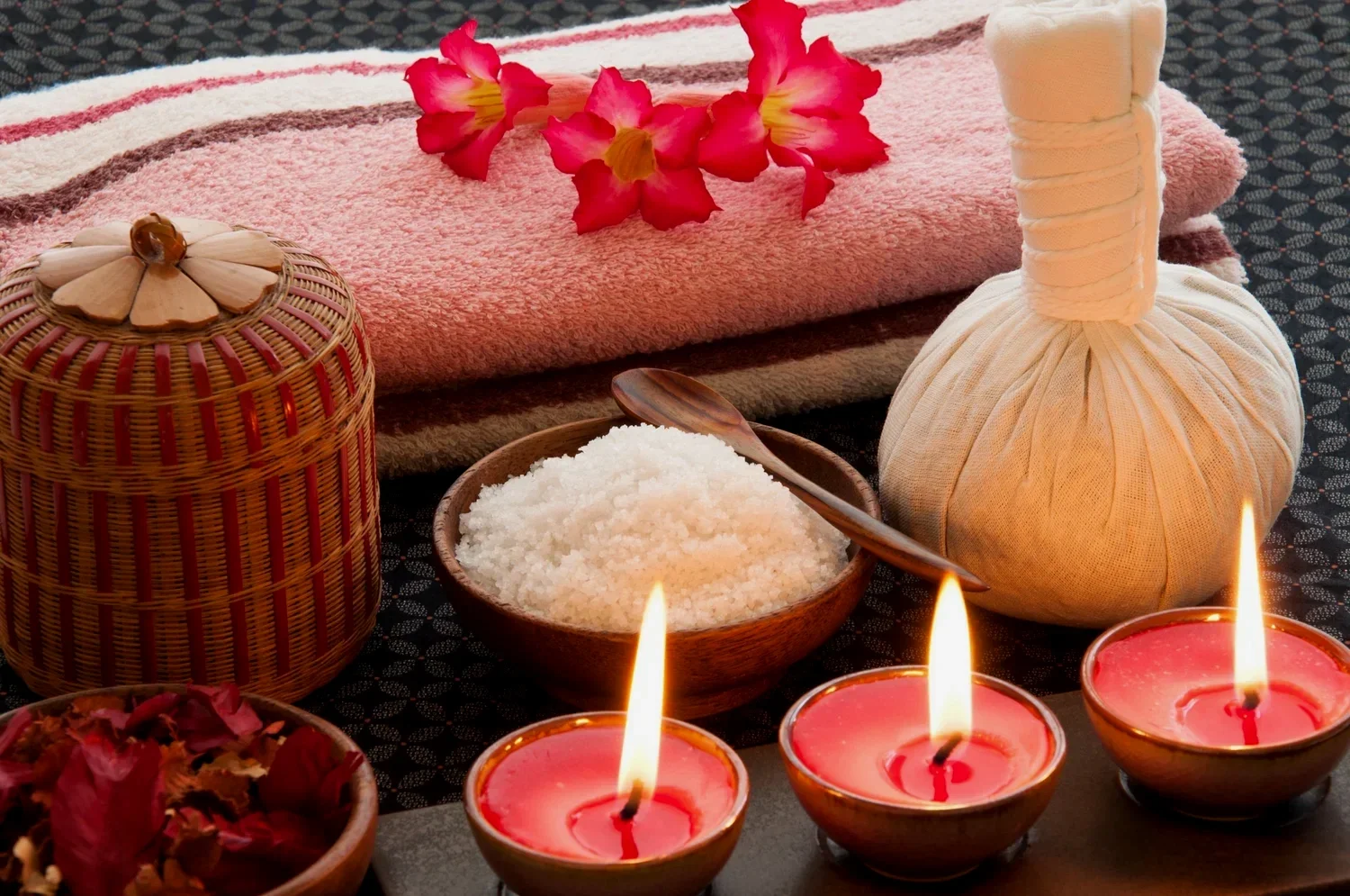 Spa setup with pink flower petals, folded towel, herbal compress ball, and candles.