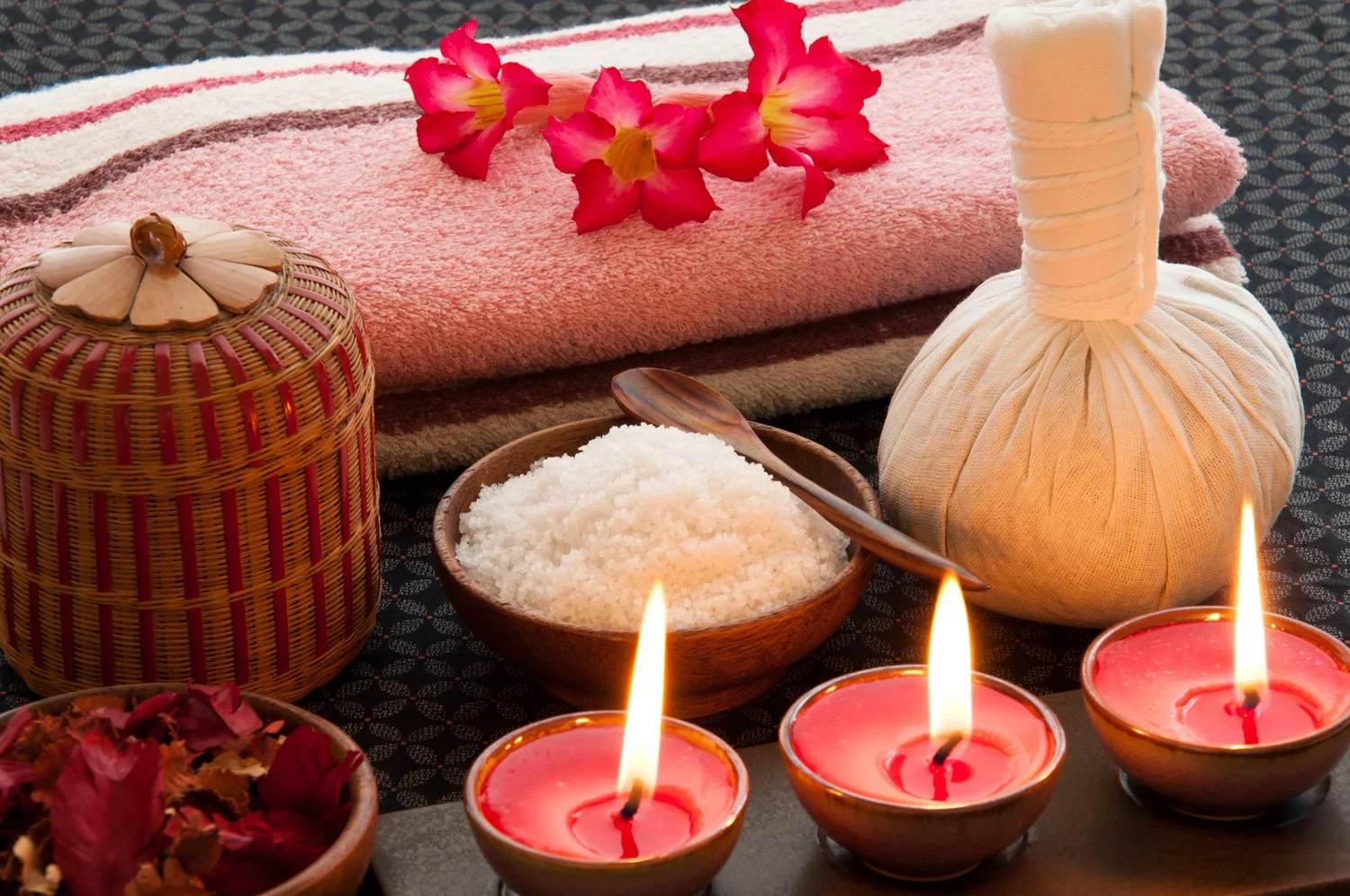 Spa setup with pink towels, pink flowers, candles, a bowl of bath salts, a herbal compress ball, and essential oils, arranged on a black patterned surface.
