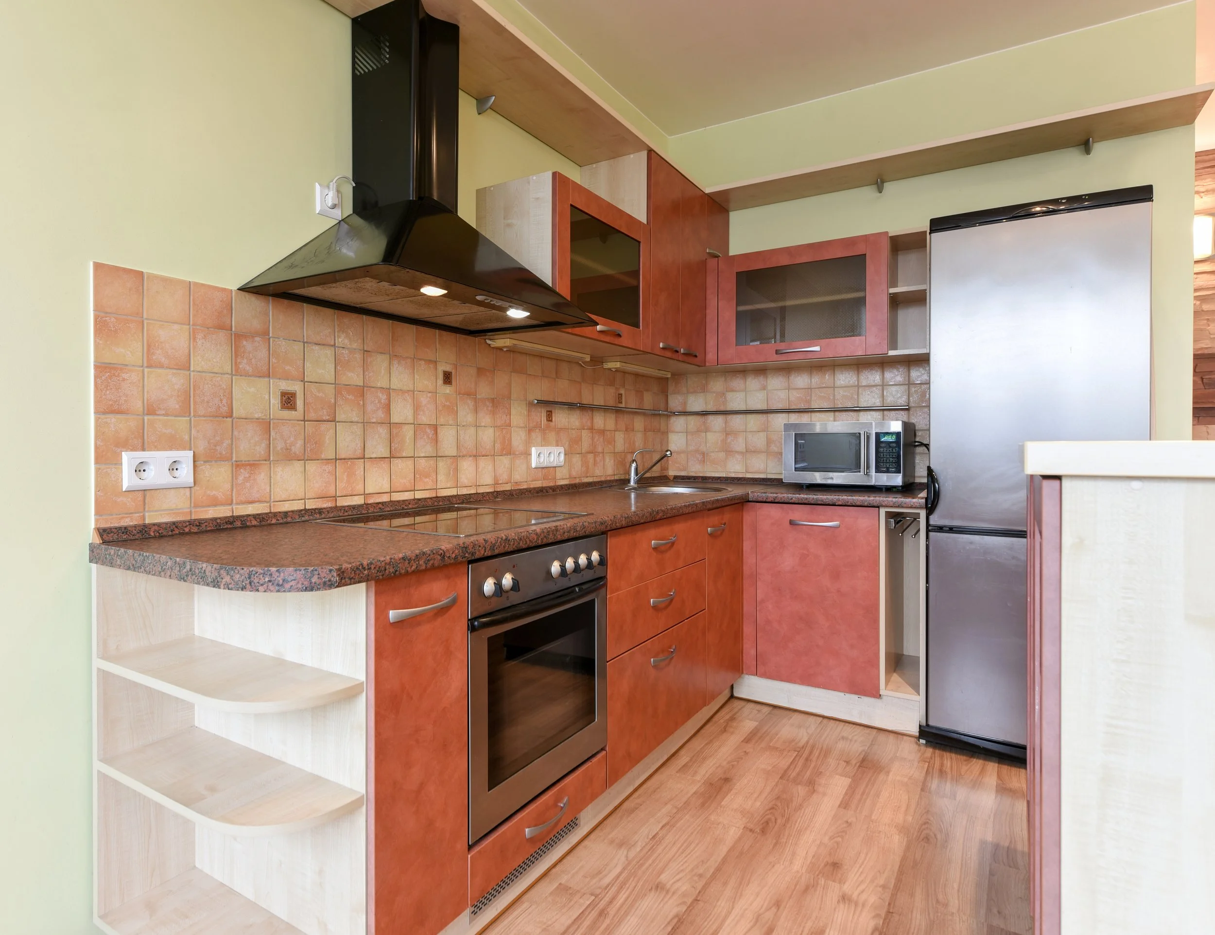 Kitchen with wooden cabinets, orange tiled backsplash, black range hood, microwave, stainless steel refrigerator, and wooden flooring.