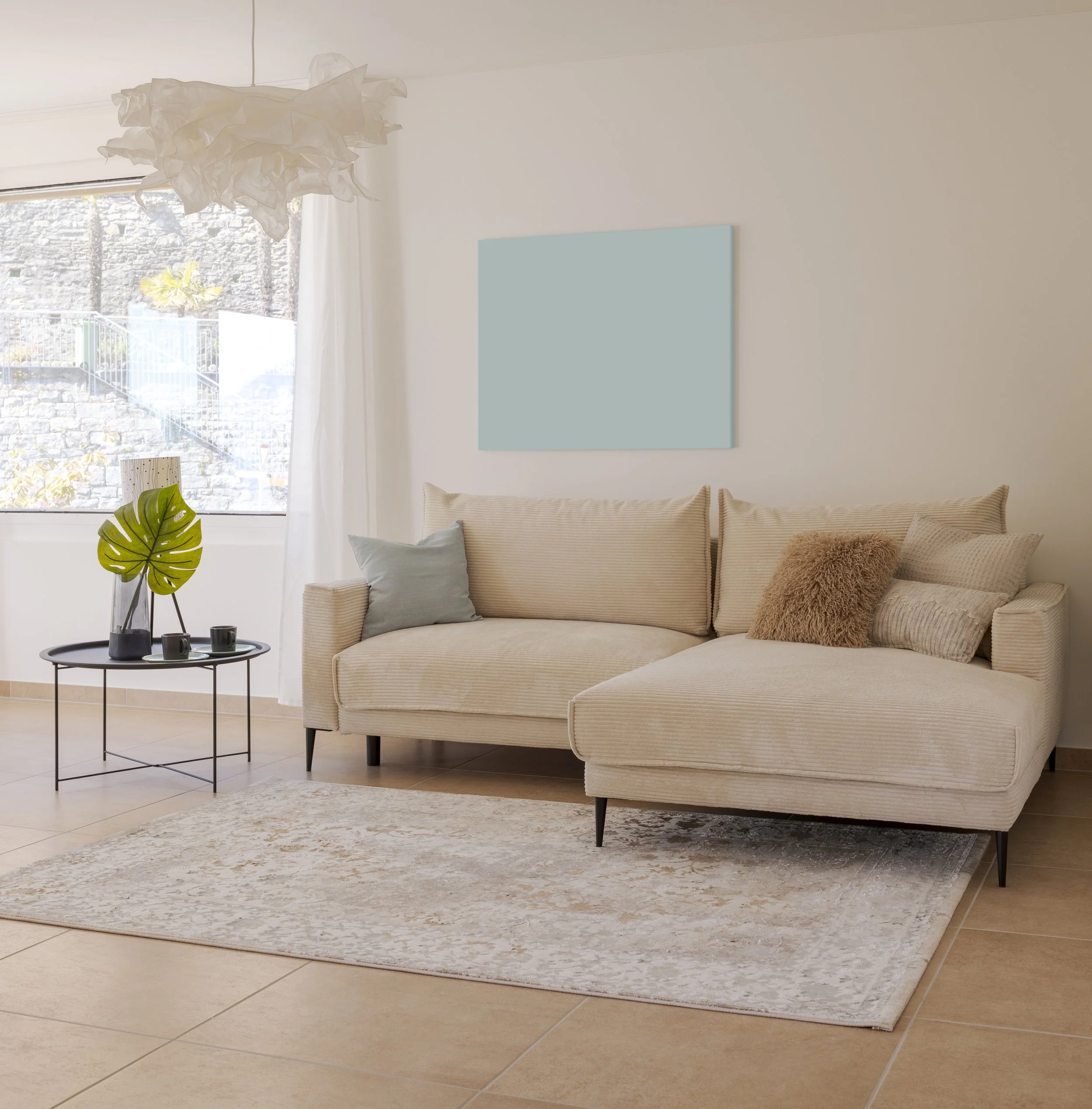 Living room with beige sofa, light blue pillow, brown furry pillow, white curtains, a round black side table with decorative items, a large window, a white paper lantern ceiling light, and a blank light blue wall art piece.