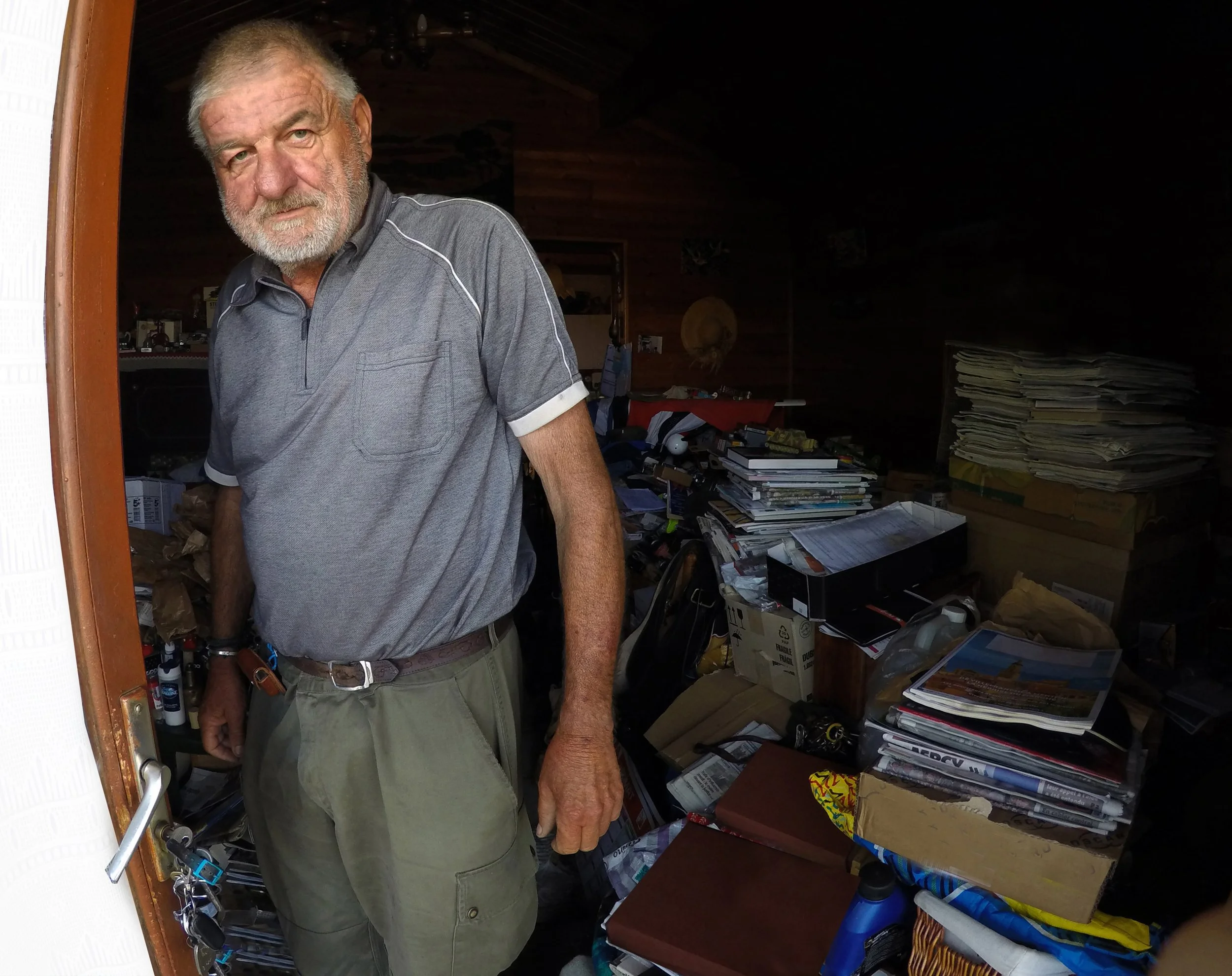 An elderly man with a beard and gray hair standing in a cluttered room filled with papers, books, and boxes.