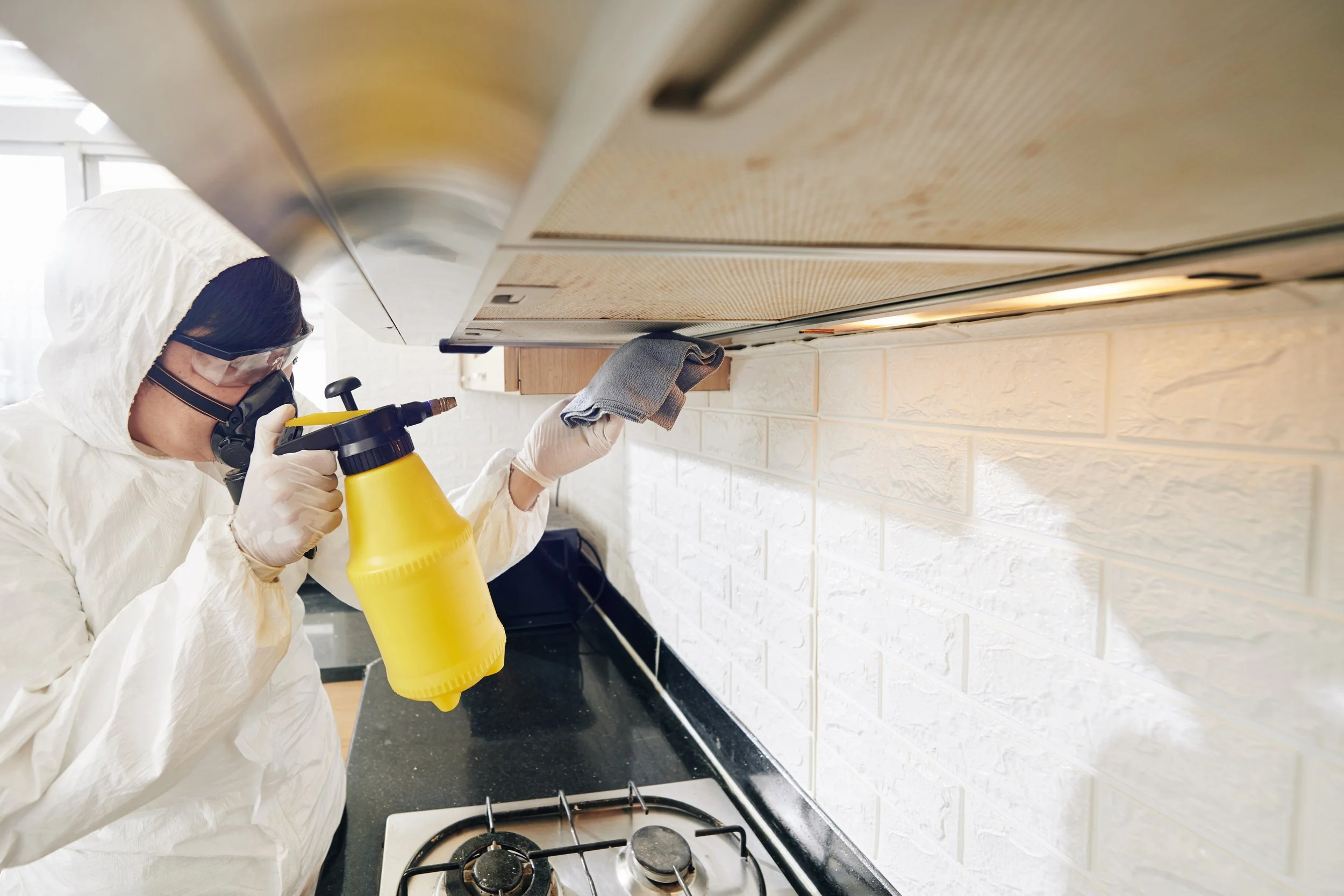 A person in protective clothing and goggles disinfects kitchen cabinets with a spray bottle.