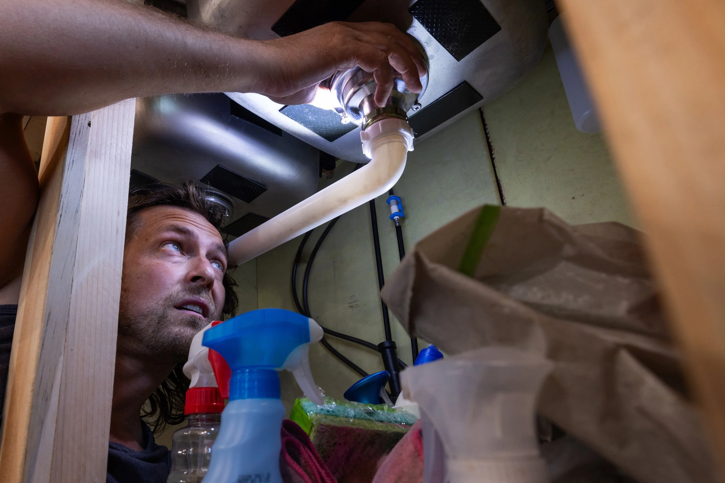 Man underneath a kitchen sink, inspecting plumbing with tools, surrounded by cleaning supplies and a paper bag.