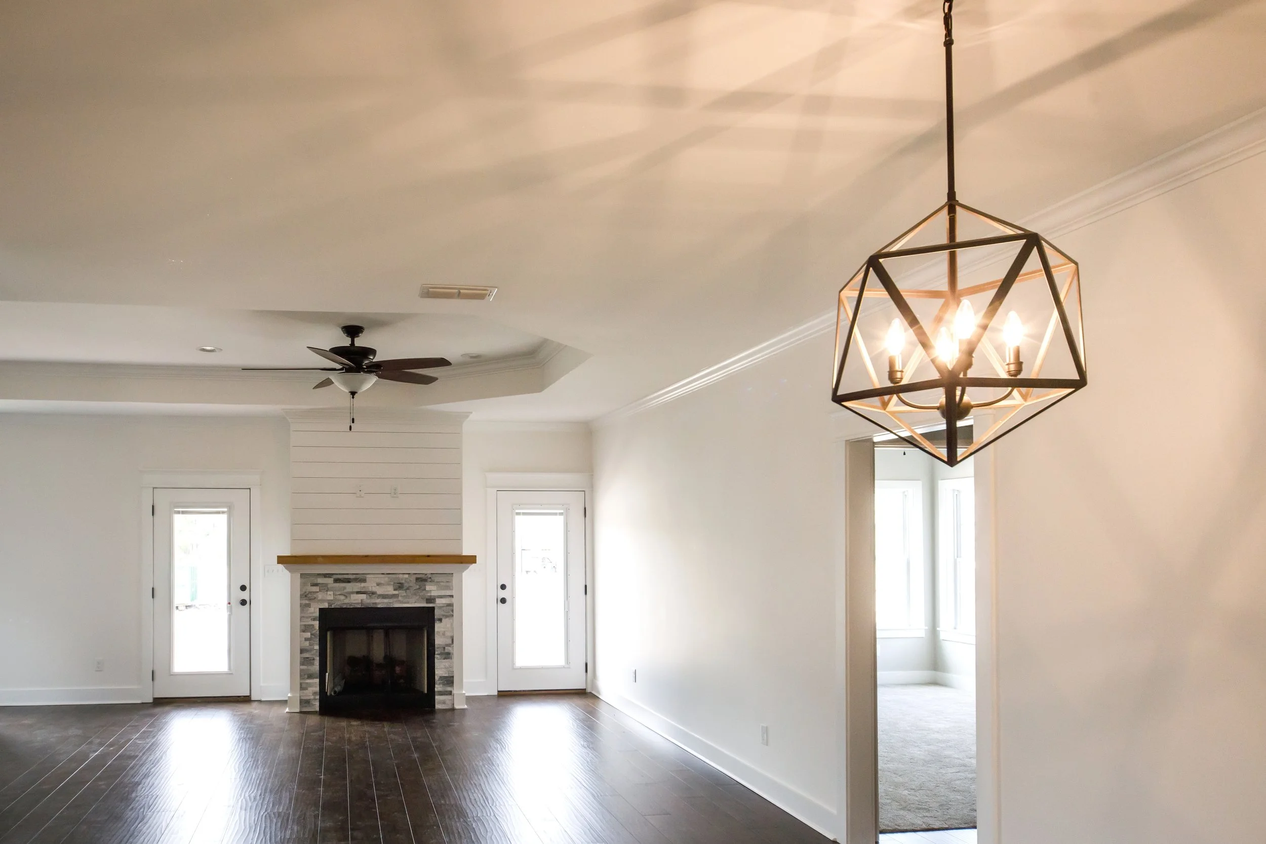 Empty living room with white walls, dark hardwood floors, a fireplace with a brick surround, a ceiling fan, and a geometric pendant light. Natural light coming through windows and external door.