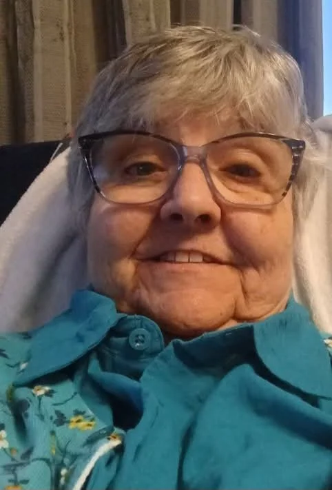 Close-up photo of an elderly woman with short gray hair, wearing glasses and a teal shirt, smiling.