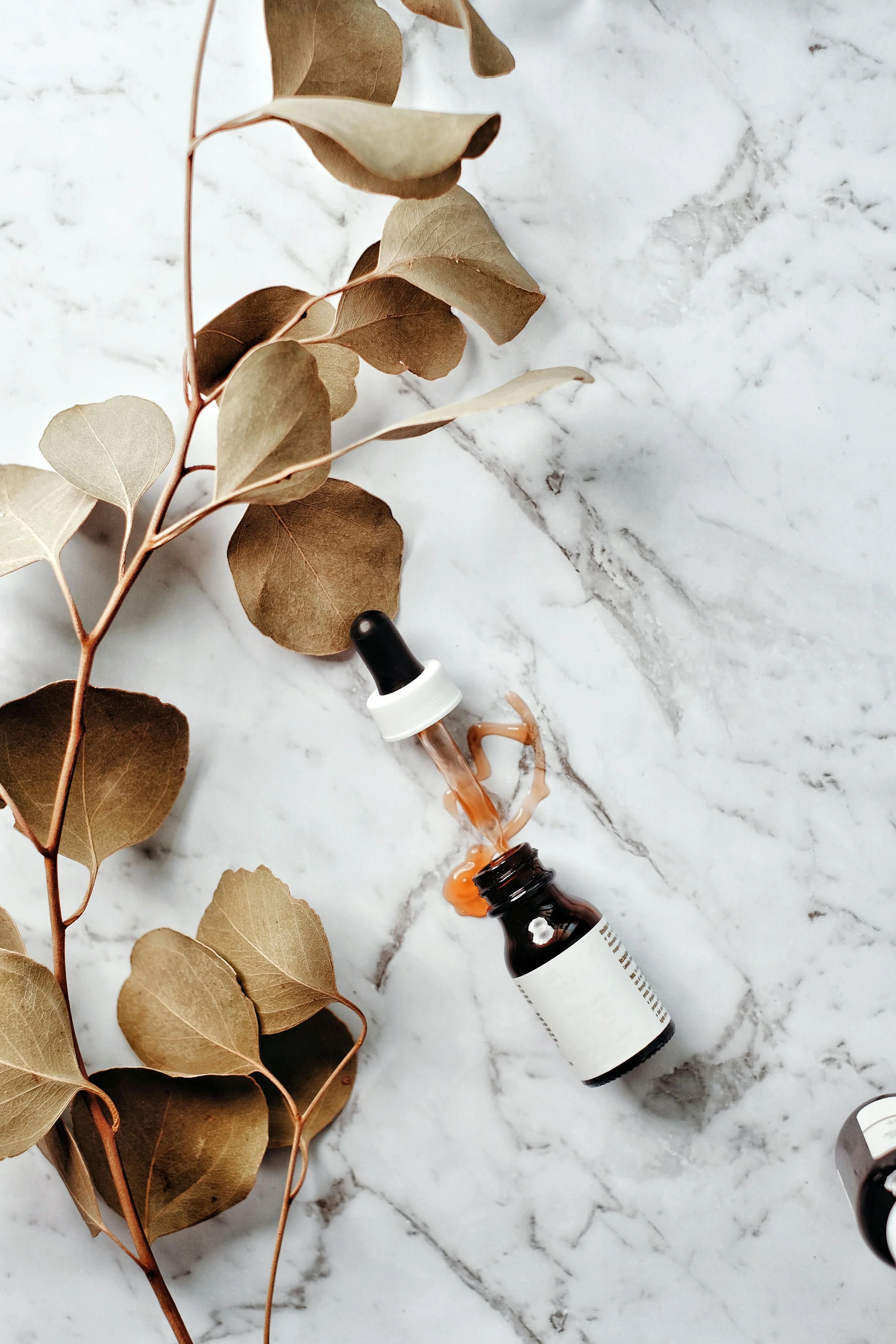 An overhead view of a brown dropper bottle with an open white cap, a few drops of liquid inside the dropper, spilled onto a white marble surface, with dried eucalyptus leaves nearby.
