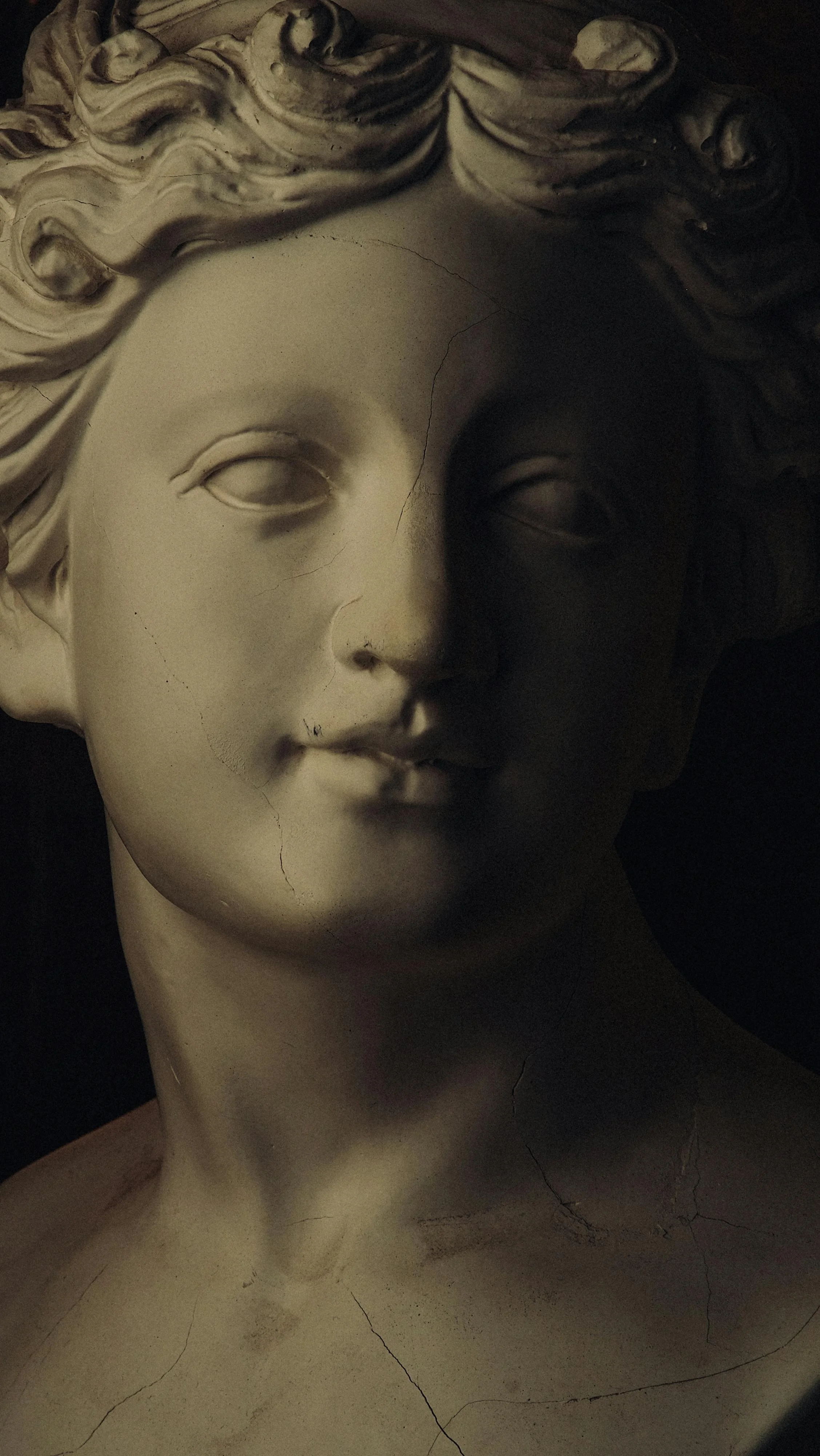 Close-up of a classical sculpture bust of a woman's face with curly hair, showing some cracks on the surface.