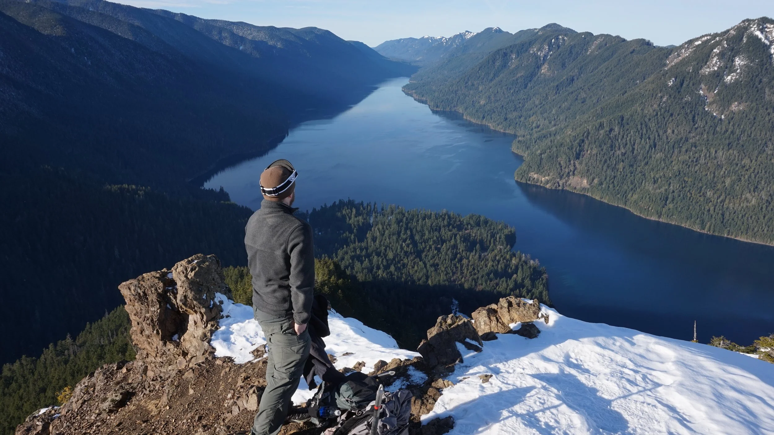 A man hiking overlooking a river and a valley far below mountains in the distance and surrounding the lake