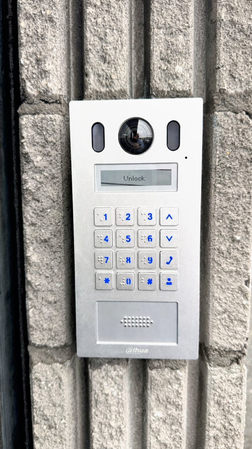 A metal intercom / access control system mounted on a brick wall. It has a camera lens, a small display screen showing 'Unlock,' and a numeric keypad with blue backlit numbers.