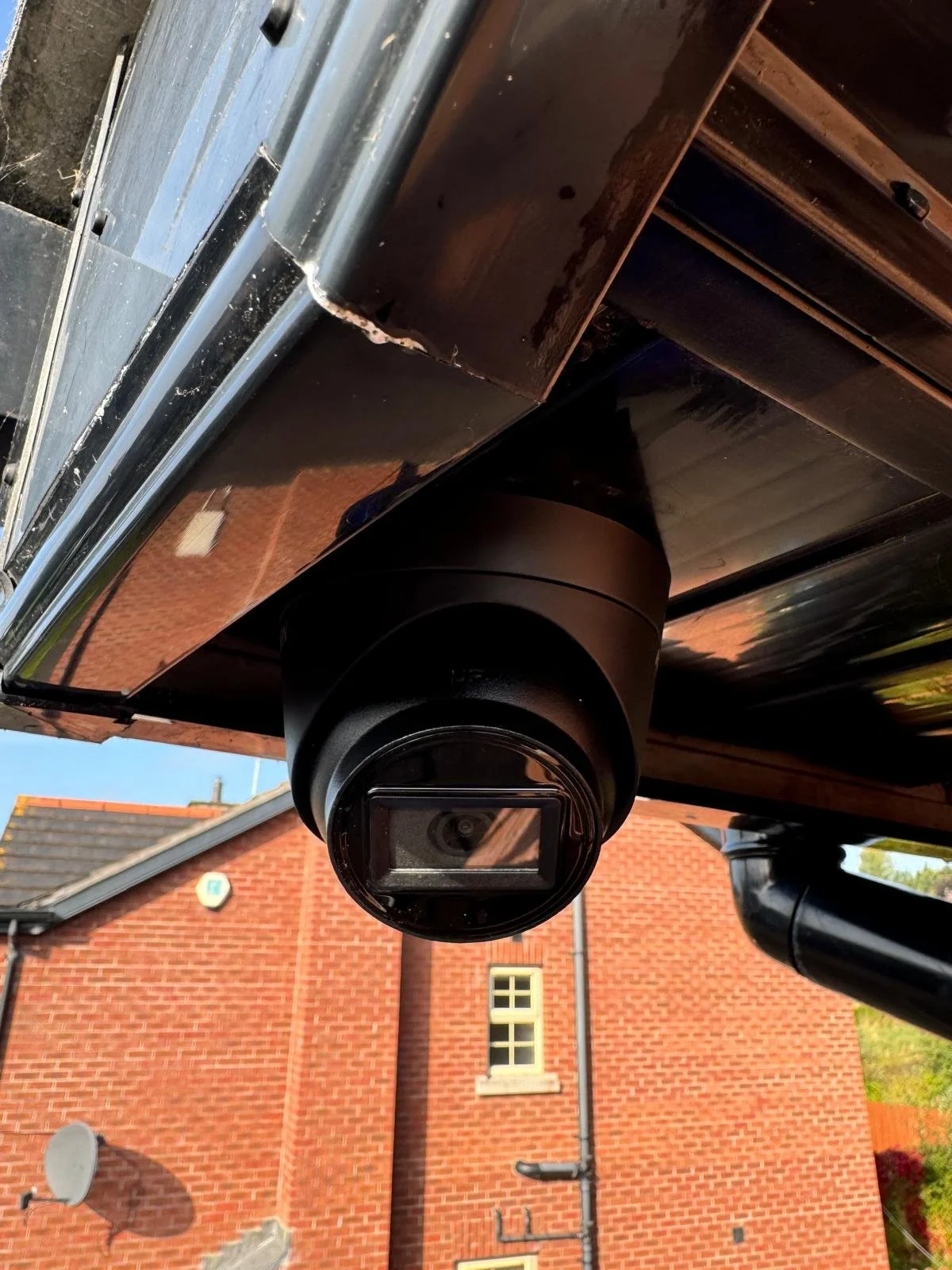 A close-up of a security camera mounted on the underside of a black surface, with a red brick building and satellite dish in the background.