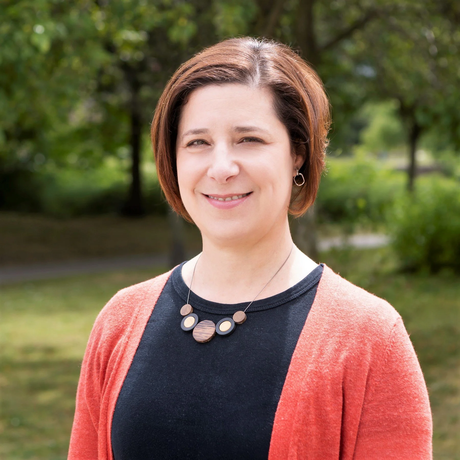 A woman with short brown hair smiling outdoors, wearing a black shirt, a coral cardigan, and a statement necklace, with greenery in the background.