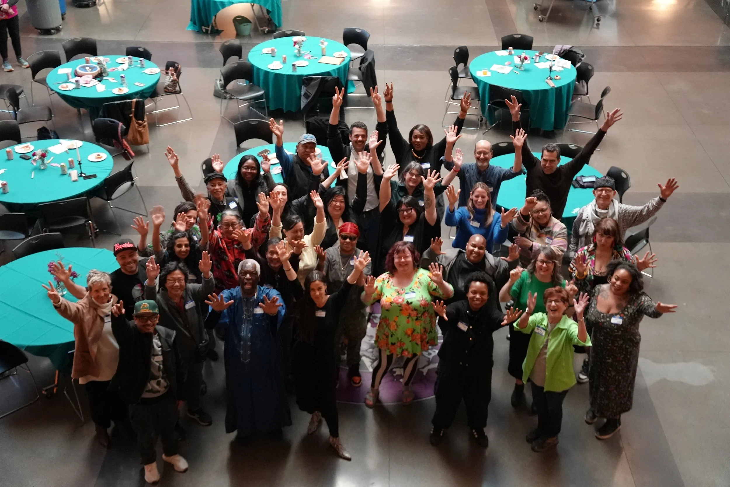 A large group of diverse people gathered together in a spacious indoor venue, smiling and raising their hands, with round tables covered with teal tablecloths and set with plates and utensils in the background.