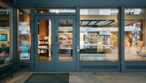 Glass storefront door and windows of a retail store with a parking lot outside.