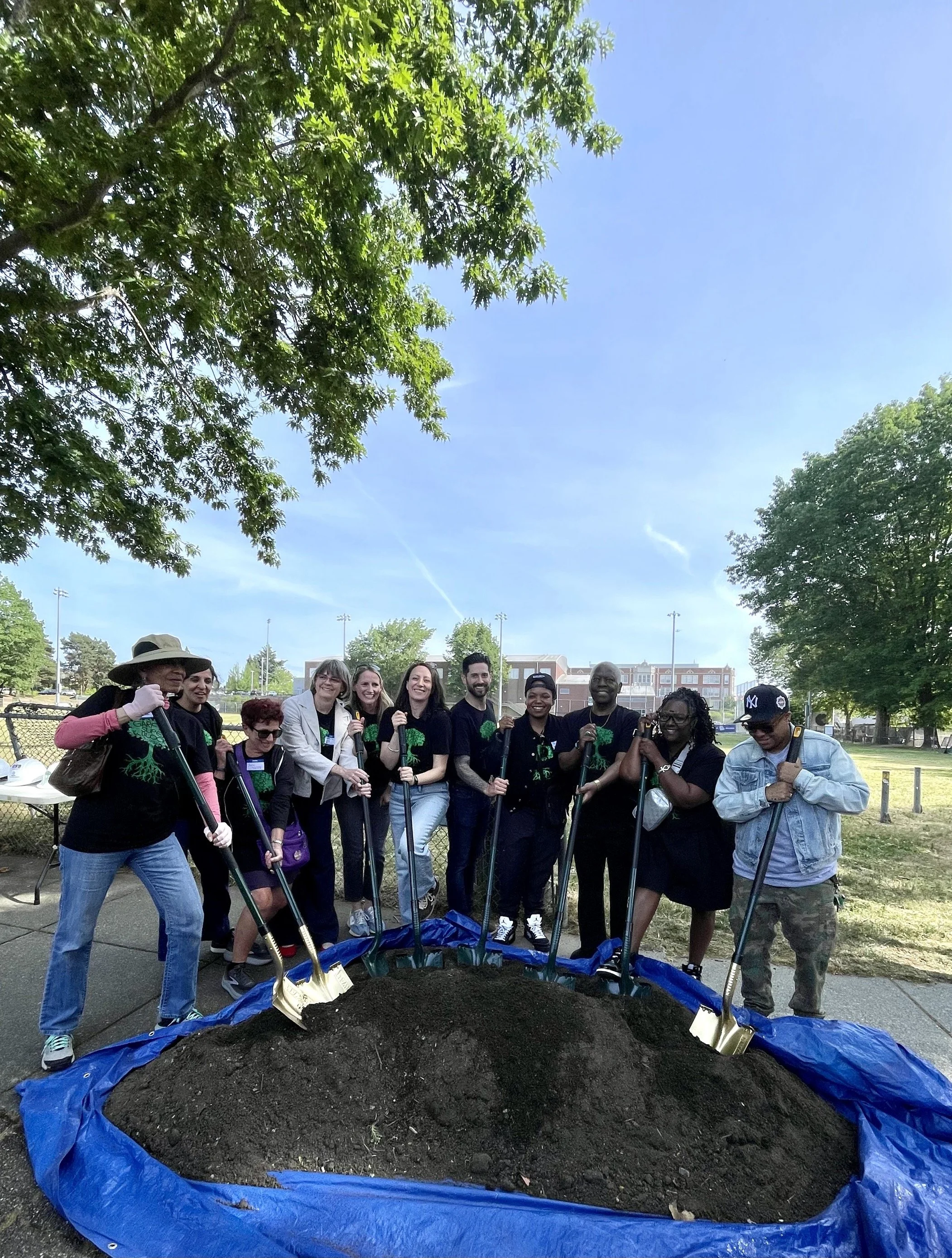 Group of people gathered outdoors for a planting event, standing around a large pile of soil with shovels, under a tree on a sunny day.