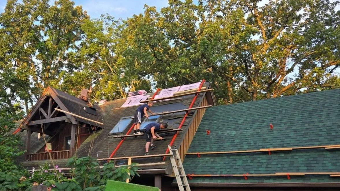 Two workers installing green roofing shingles on a house in a wooded area, with scaffolding and insulation visible.