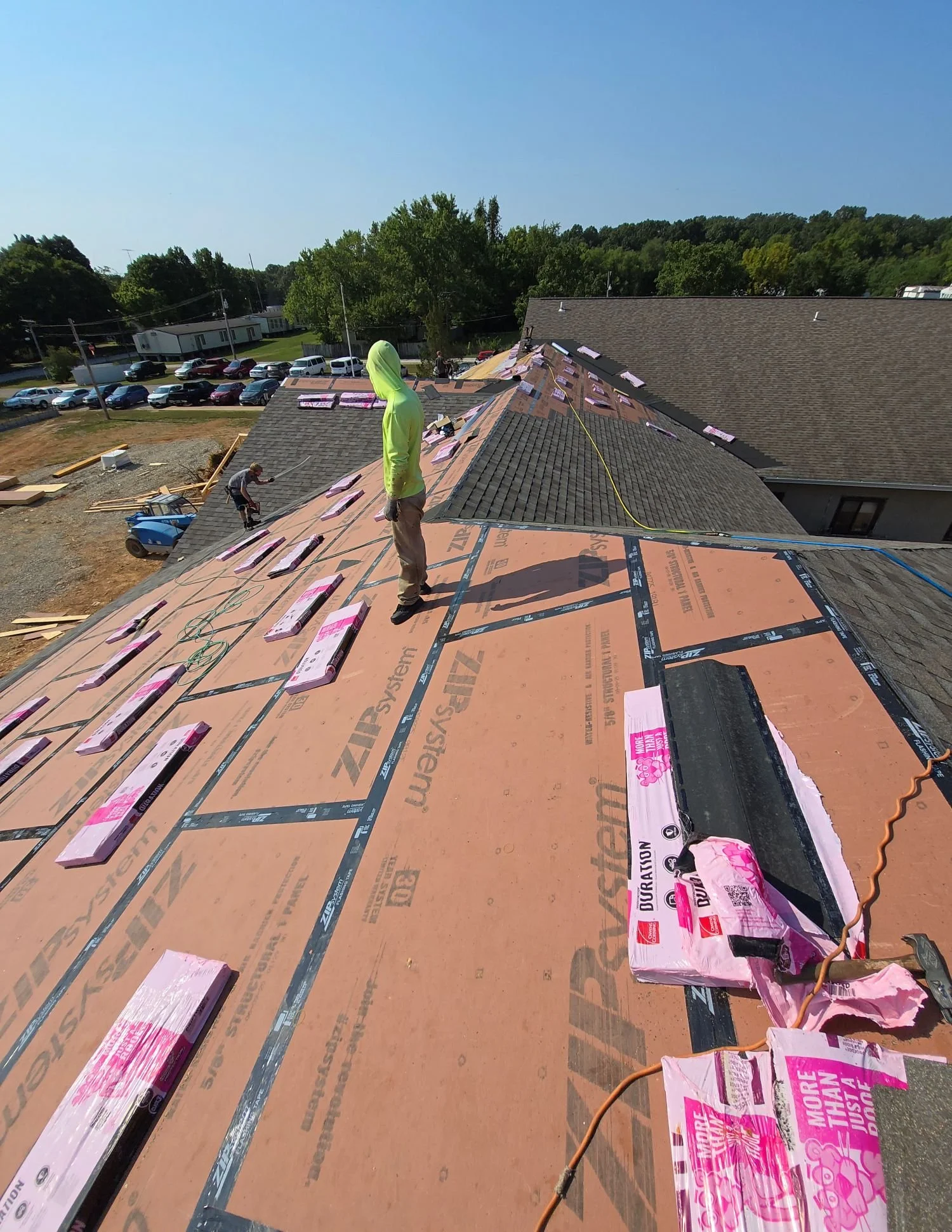 Roof under construction with pink insulation boards, workers installing new roofing material, and a person in a bright green hoodie observing. Construction tools and materials are visible, with a parking lot and trees in the background.