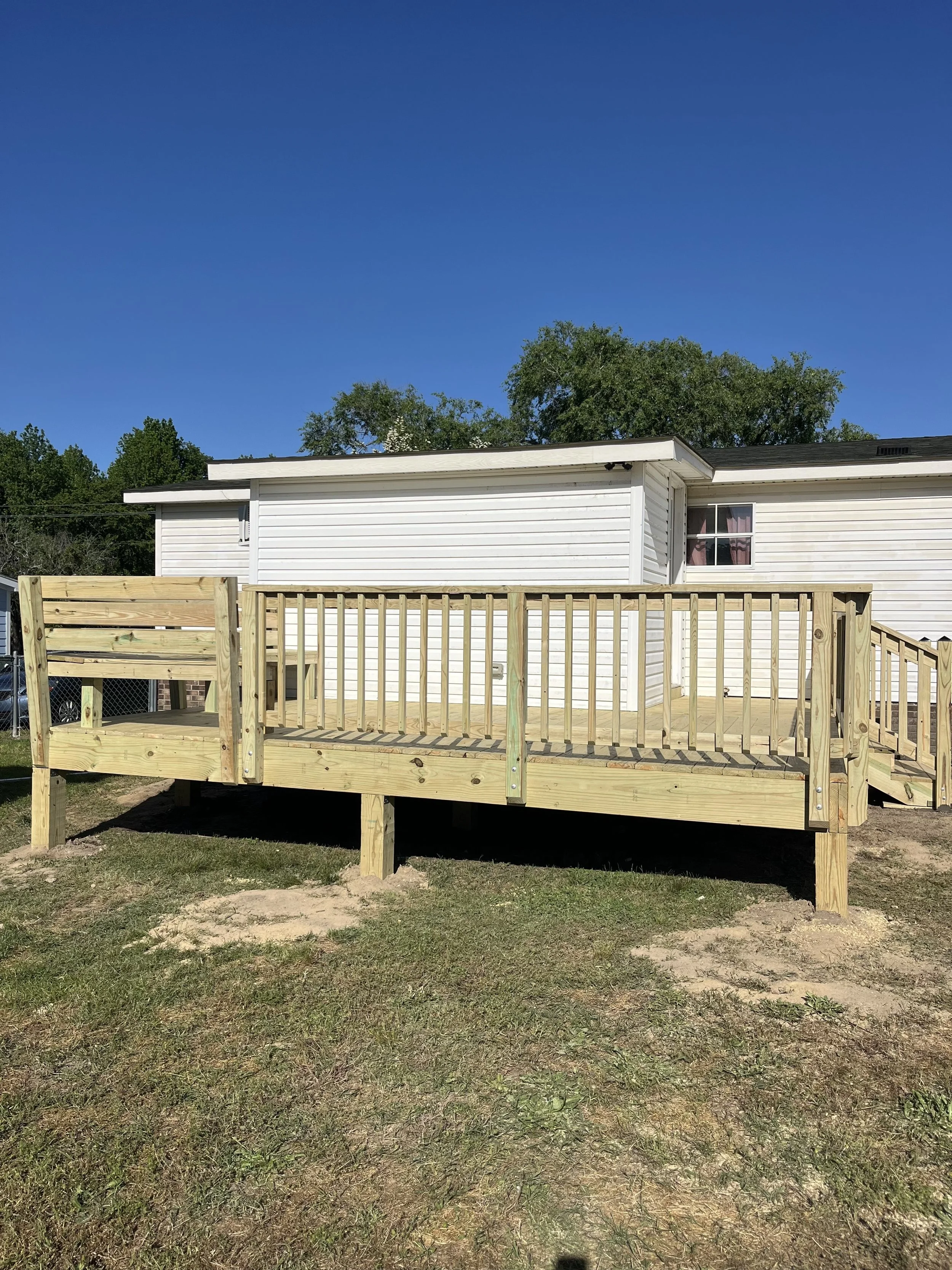 New wooden deck with railing attached to a white house, set on a grassy yard under a blue sky.