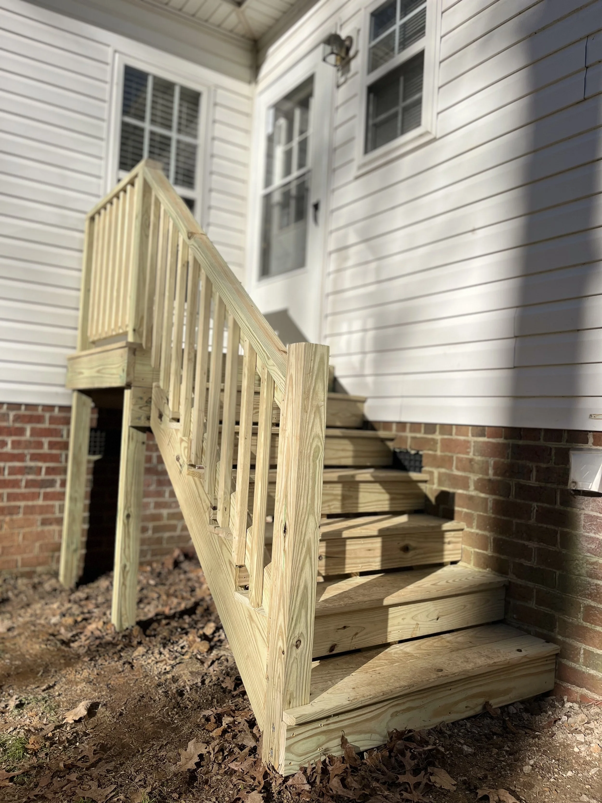New wooden staircase outside a house with white siding and brick foundation, featuring a railing on one side and a light fixture above the door.