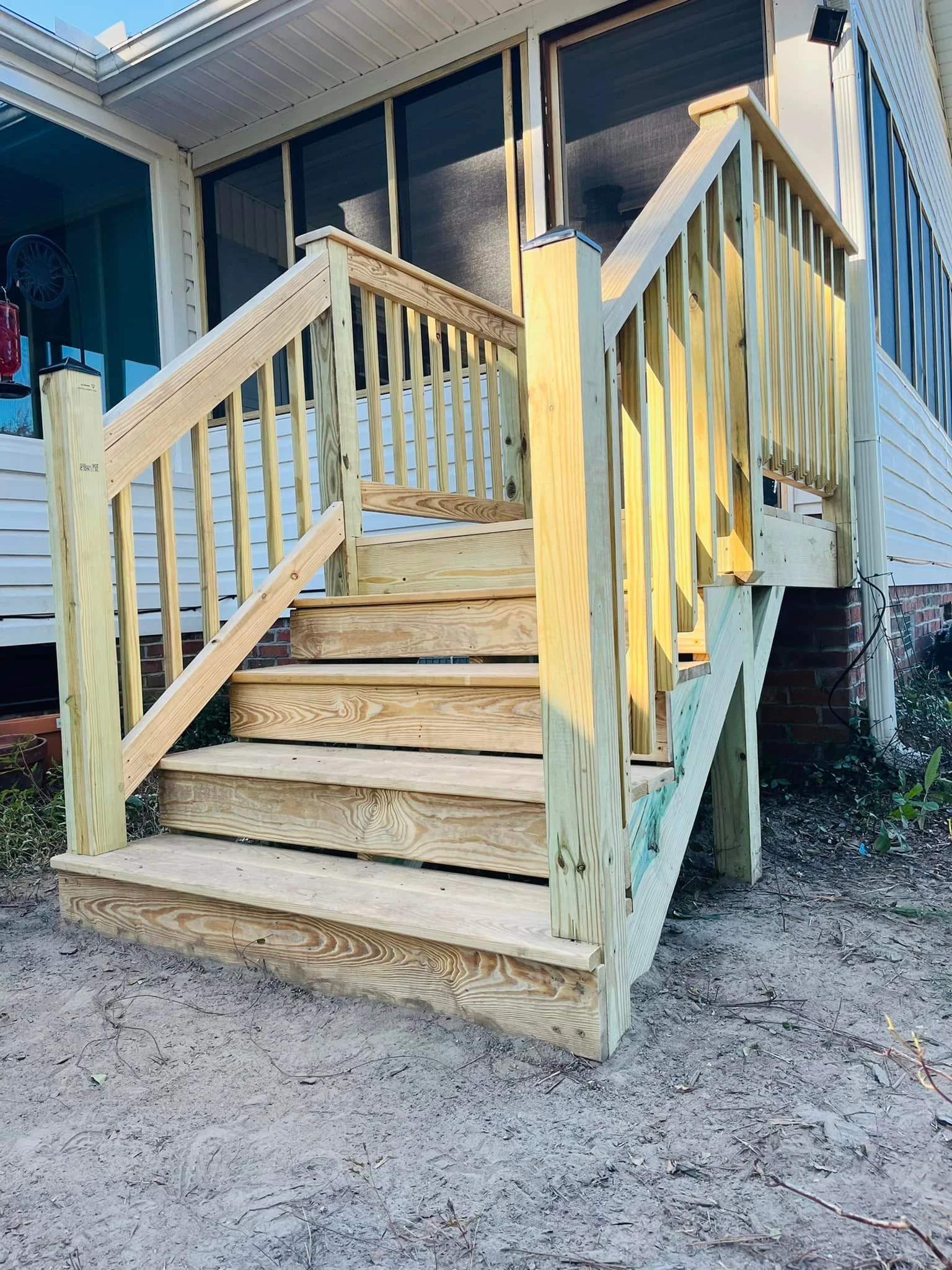 Newly built wooden deck stairs with railings attached to the house, leading up to a screened porch, with a dirt ground underneath.