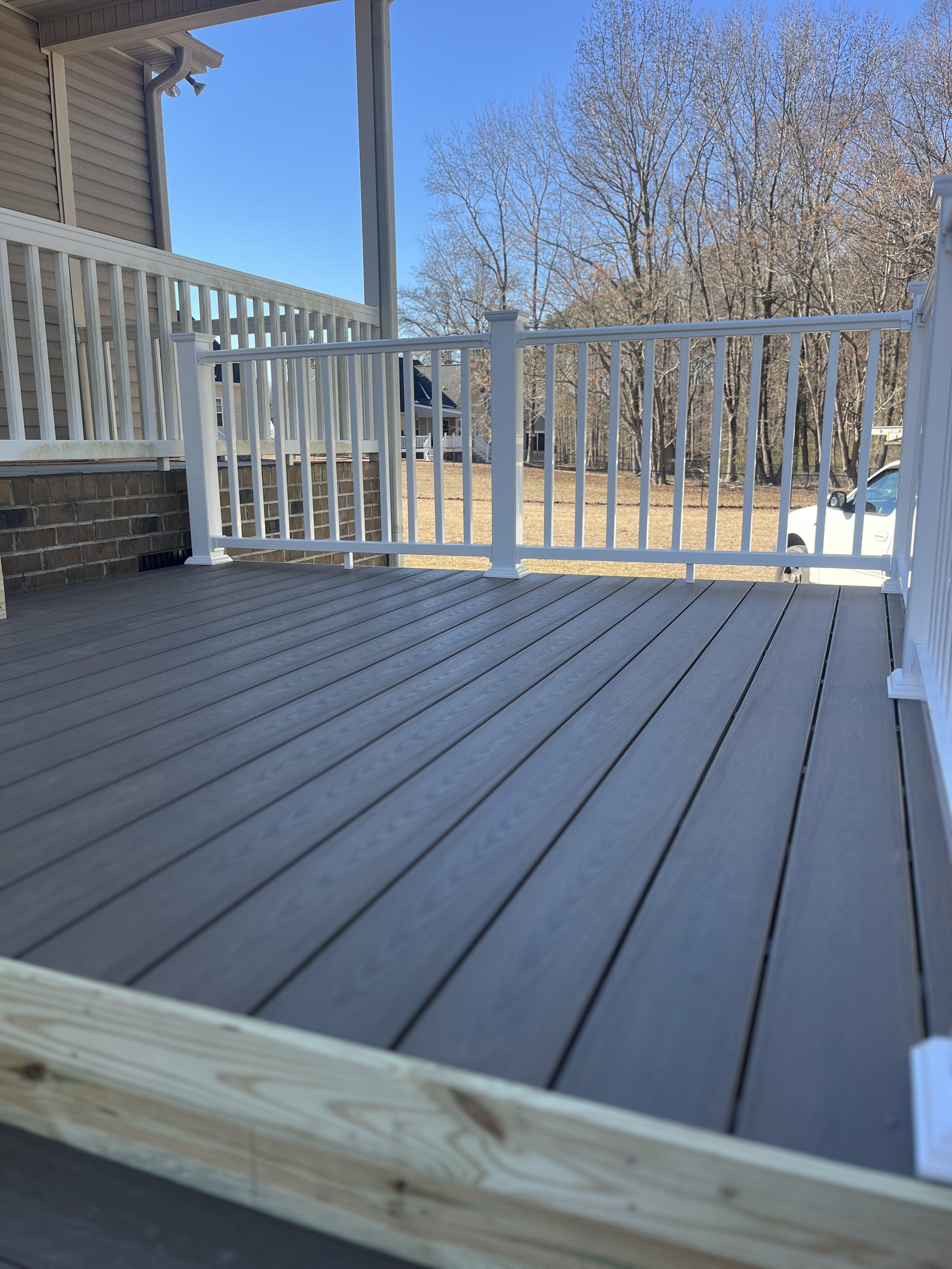 Newly built outdoor deck with gray wooden flooring and white railing, attached to a house with beige siding, in a suburban neighborhood with leafless trees and a blue sky.