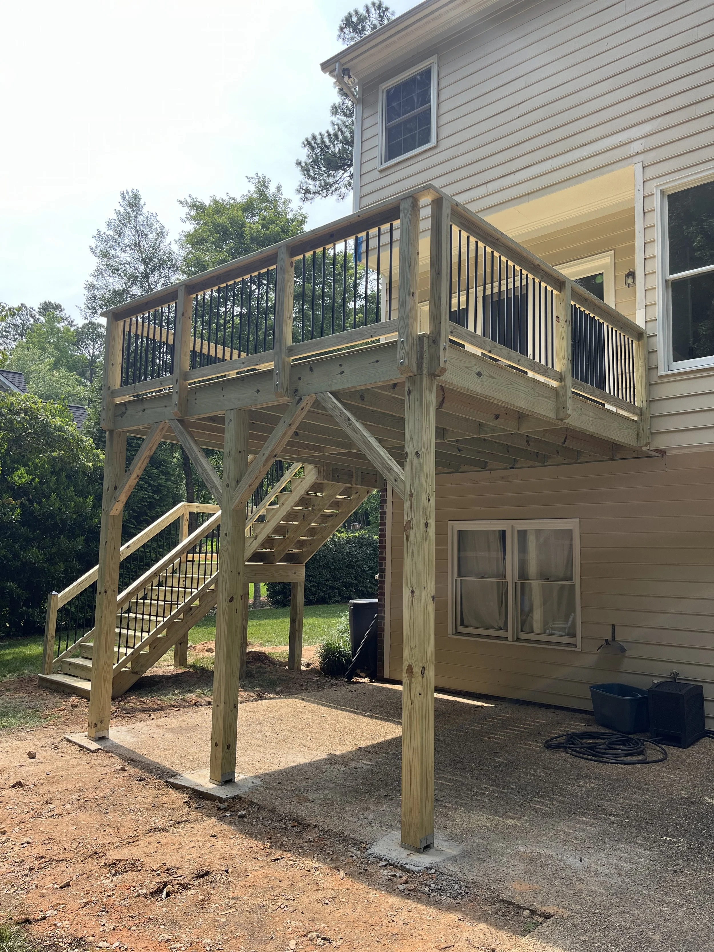 New wooden deck with staircase attached to the back of a beige house, with black metal railing and trees in the background.