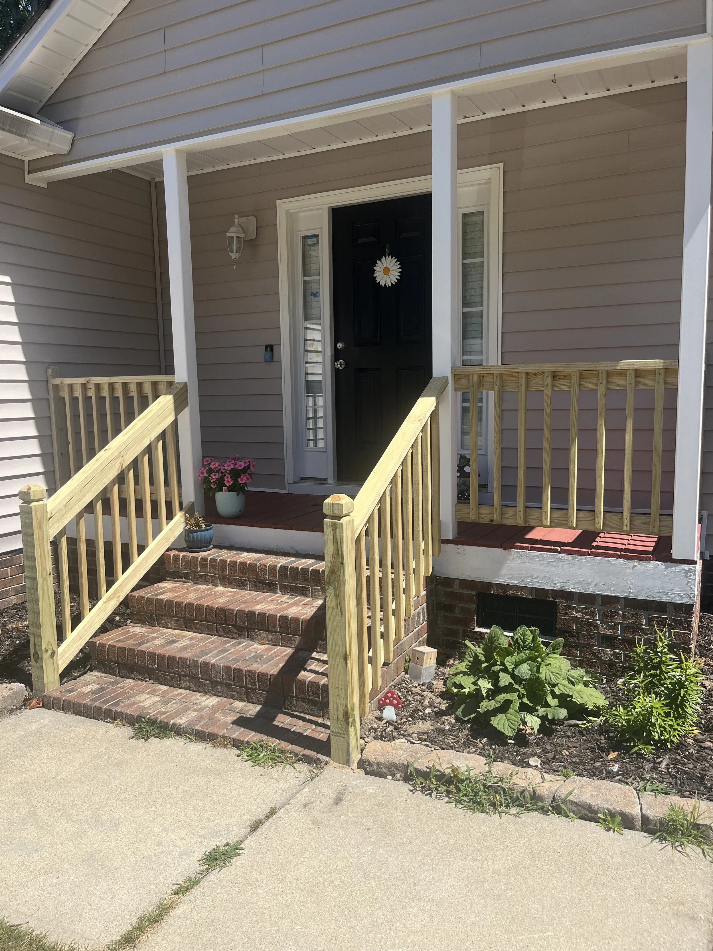 Front porch of a house with brick stairs, a small flower pot with pink flowers, a black front door with a daisy wreath, and a small garden bed with green plants and a decorative red mushroom ornament.