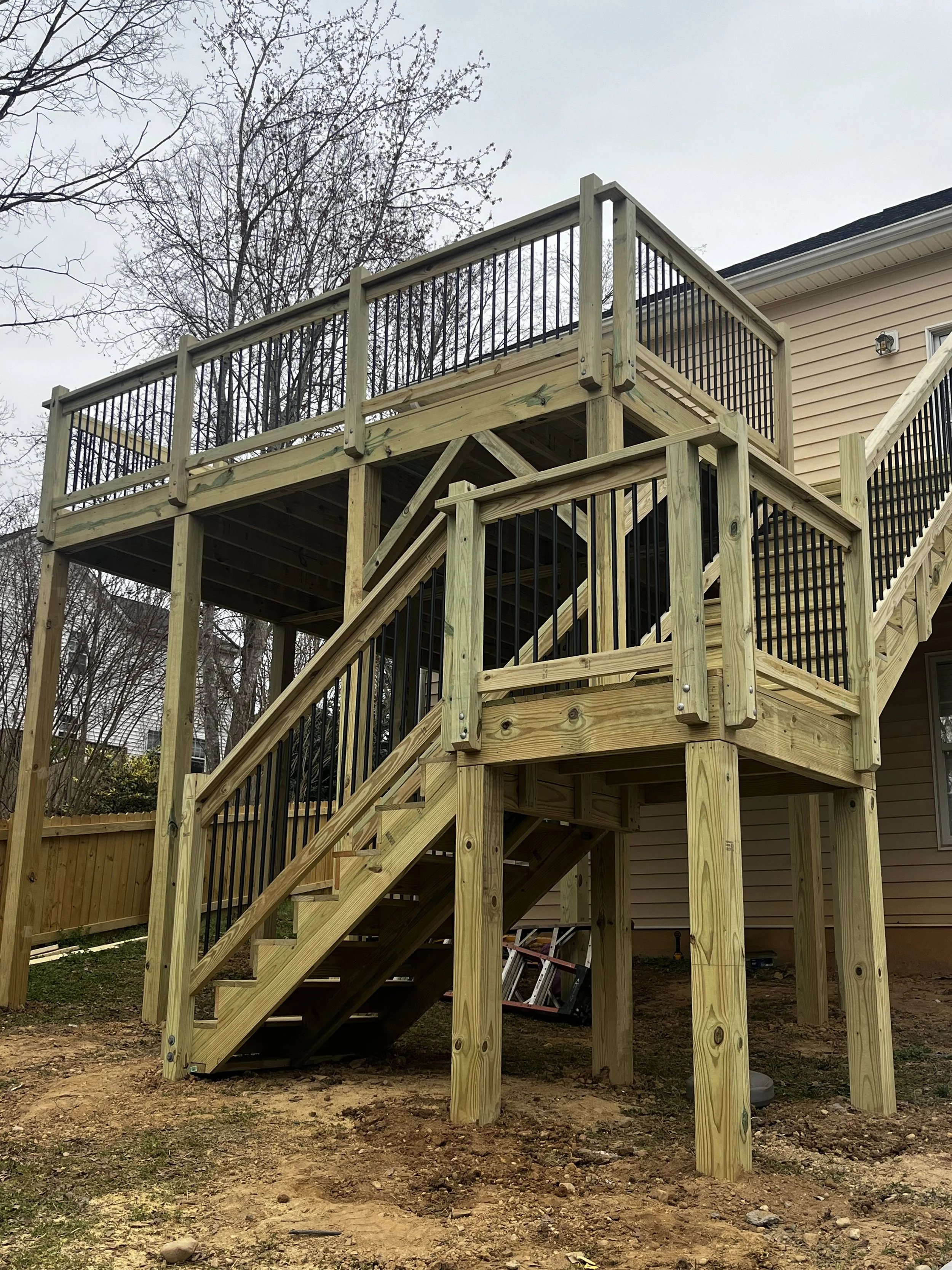 Newly built wooden deck with stairs and black metal railings in backyard next to beige house, with trees and cloudy sky in background.