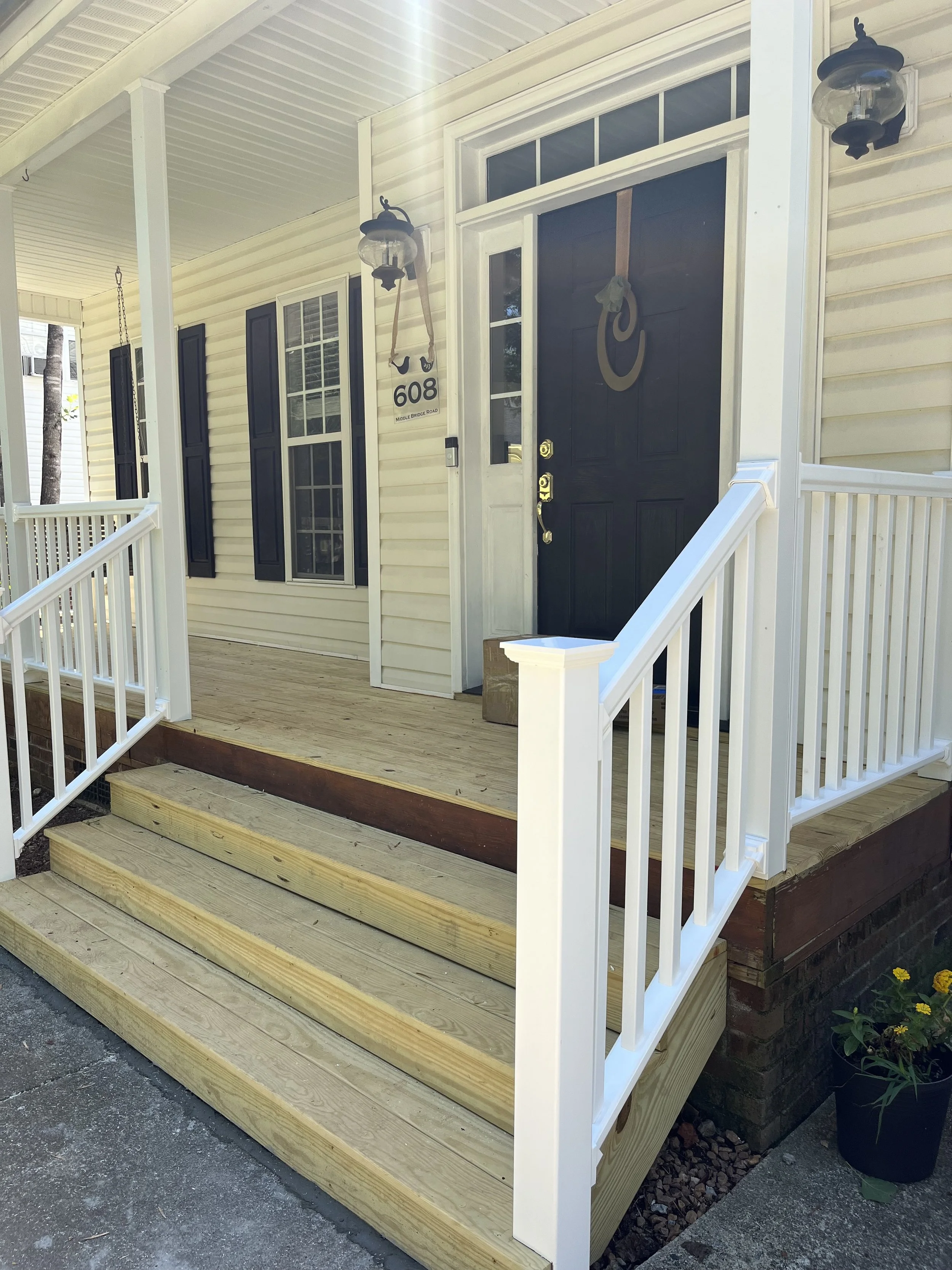 Front porch with new wooden steps and white railing, black door with a cowboy hat and ropes as decorations, house number 608, two wall lanterns, flower pot with yellow flowers beside the stairs.