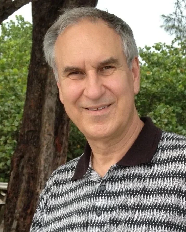 A middle-aged man with gray hair smiling outdoors with green trees in the background.