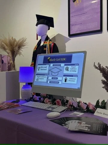 A desk setup with an informational display about Black Girl EDU, decorated with pink flowers, and a mannequin dressed in graduation attire in the background. There is a framed poster on the wall, a lamp, and some informational pamphlets on the table.
