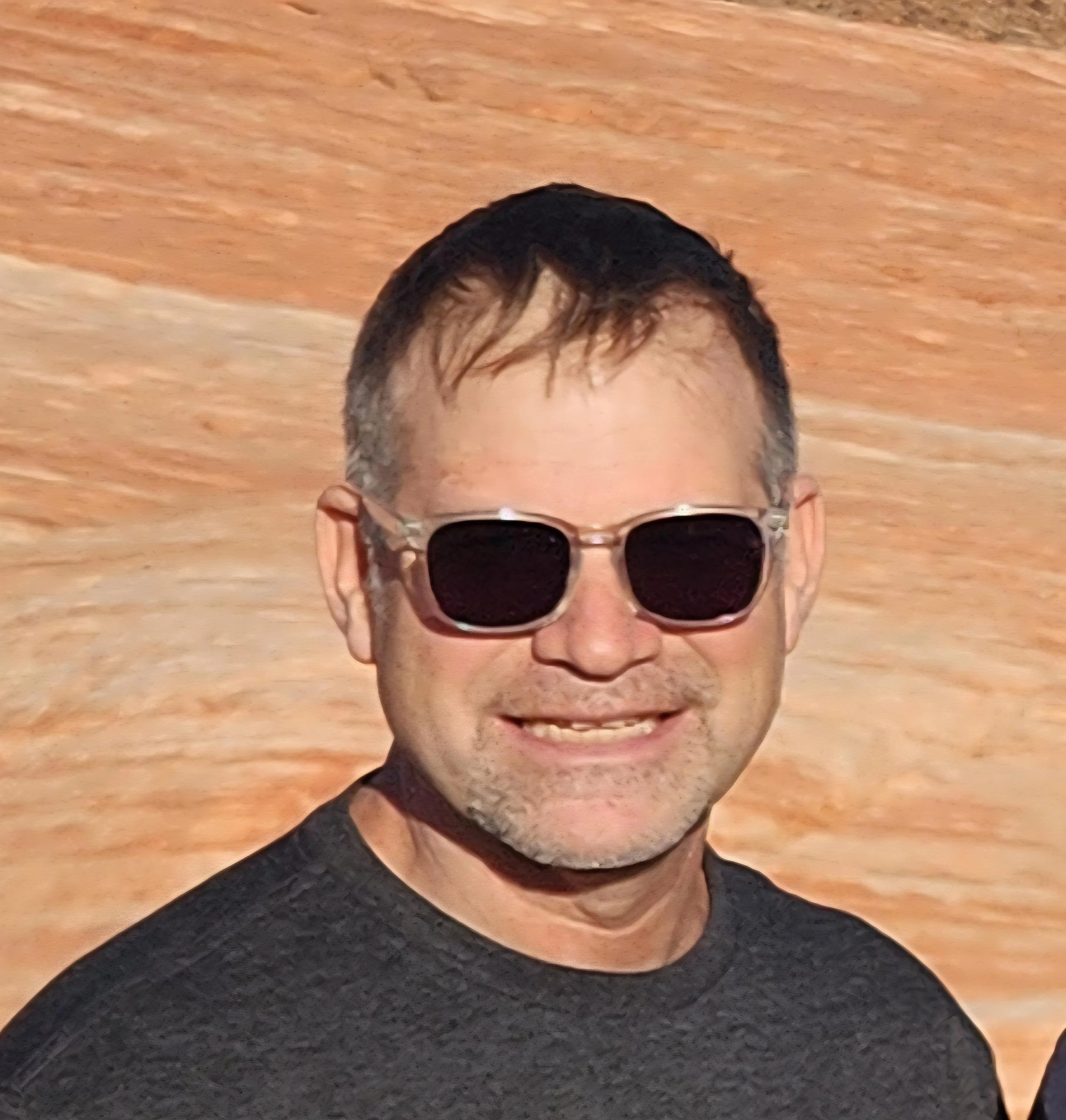 Man smiling wearing sunglasses and a black t-shirt, with a sandy colored background.