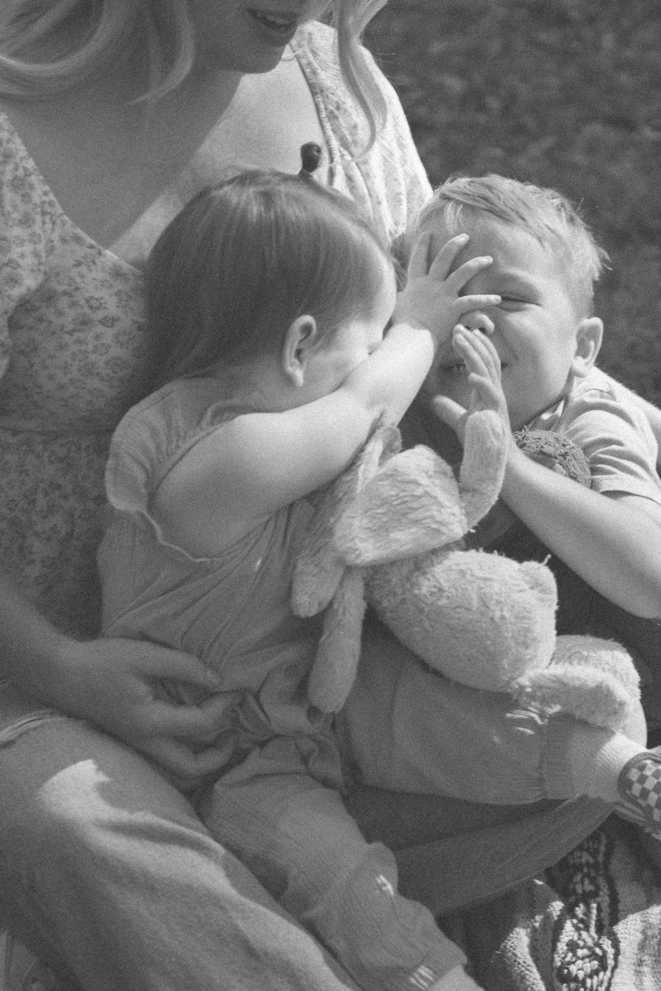 Two young children and an adult female playing with a stuffed bunny outdoors. The girl is covering the boy's face with her hand, and they appear to be laughing and having fun.