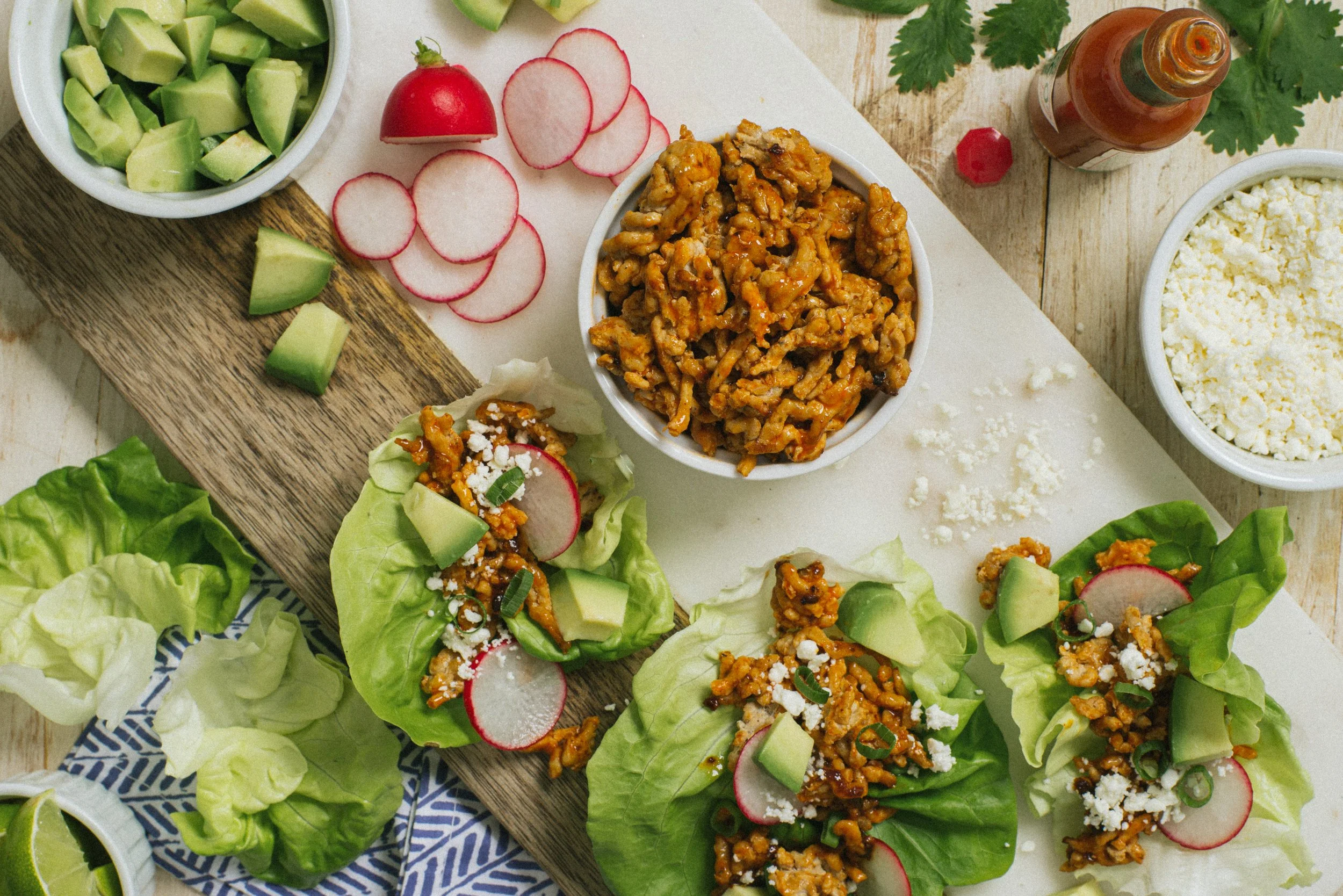 Lettuce wraps filled with seasoned ground meat, sliced radishes, diced avocado, crumbled cheese, and chopped green onions, with bowls of chopped cucumbers, crumbled cheese, hot sauce, and lime slices on a wooden and marble surface.