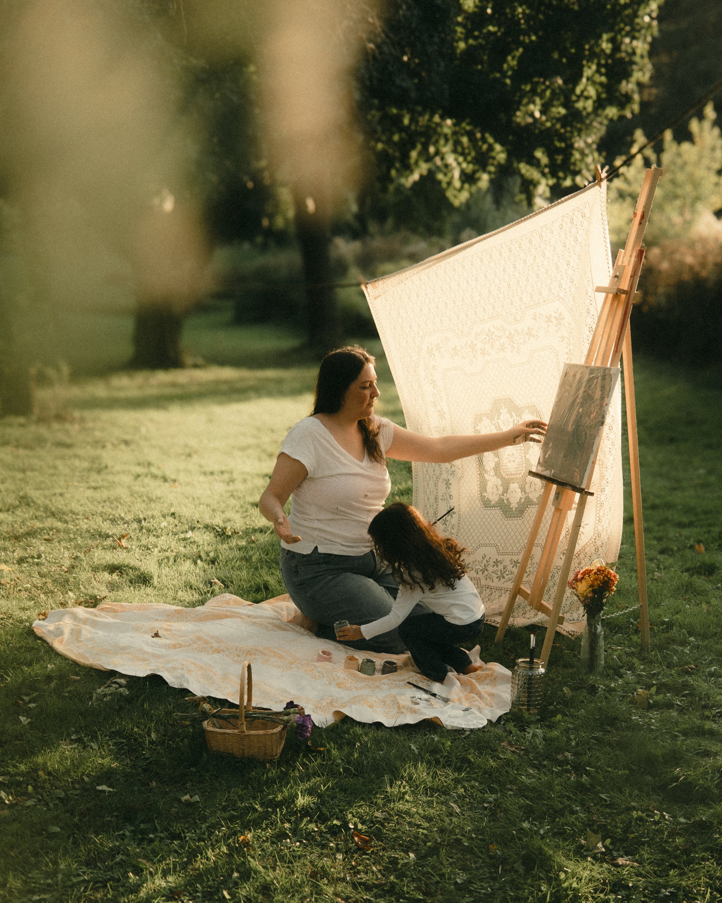 A woman and a young girl sitting on a blanket outdoors, painting on a large canvas that is propped up against an easel. The scene is set in a grassy area with trees in the background, and sunlight filtering through the leaves.