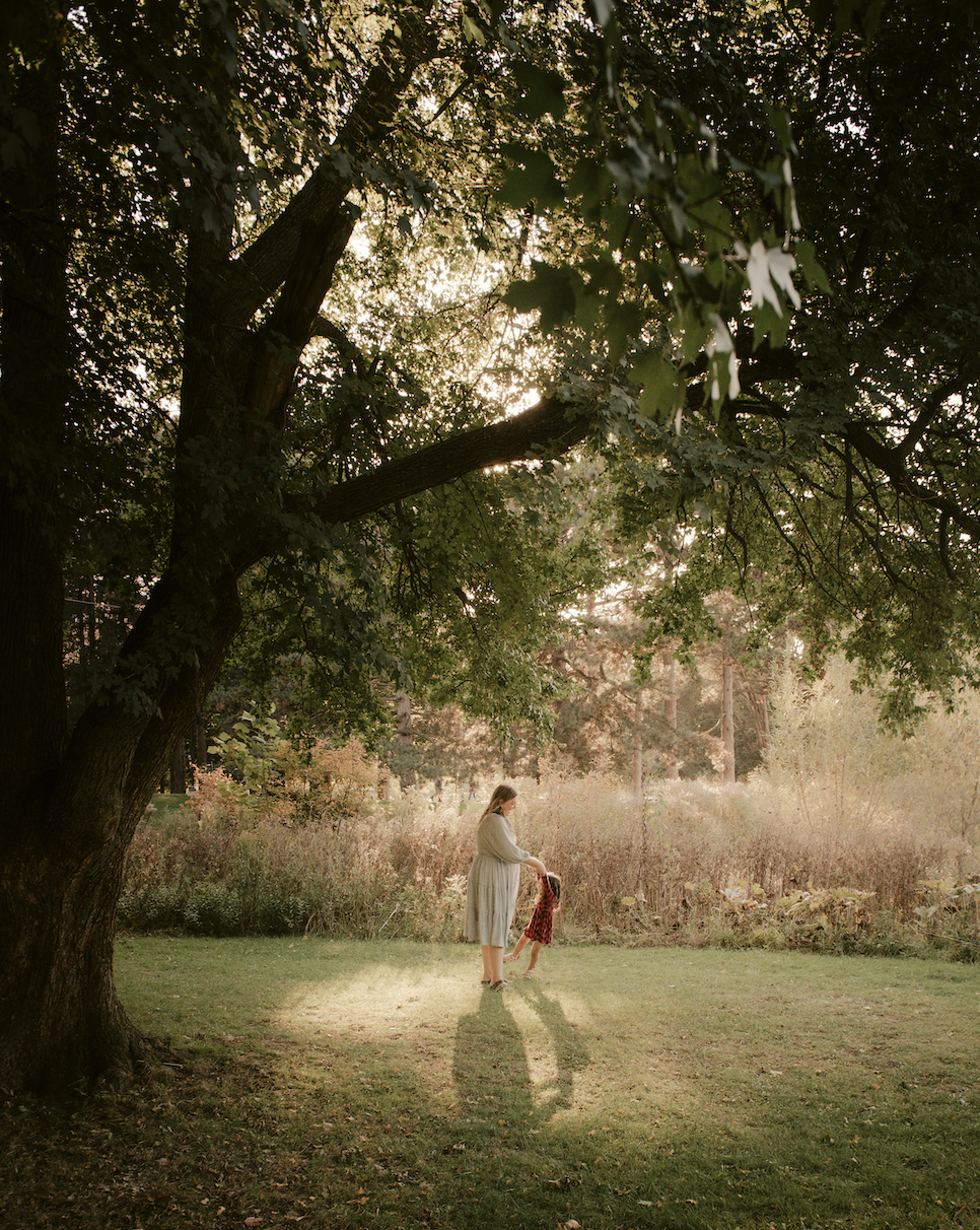 A woman and a girl holding hands in a grassy field with tall trees and sunlight filtering through the leaves.