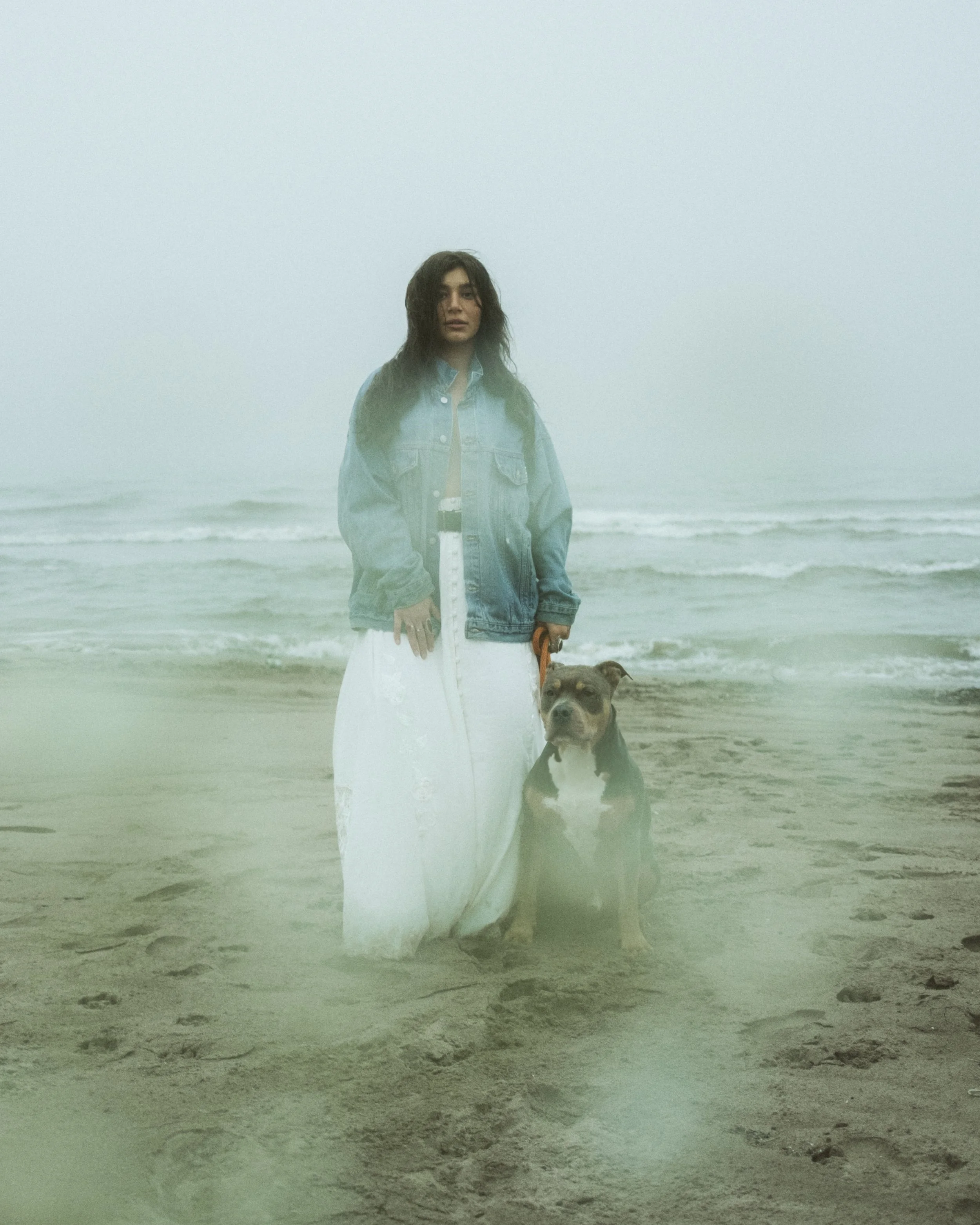 A woman standing on the beach with her dog, foggy atmosphere, in casual clothing including a denim jacket and white dress.