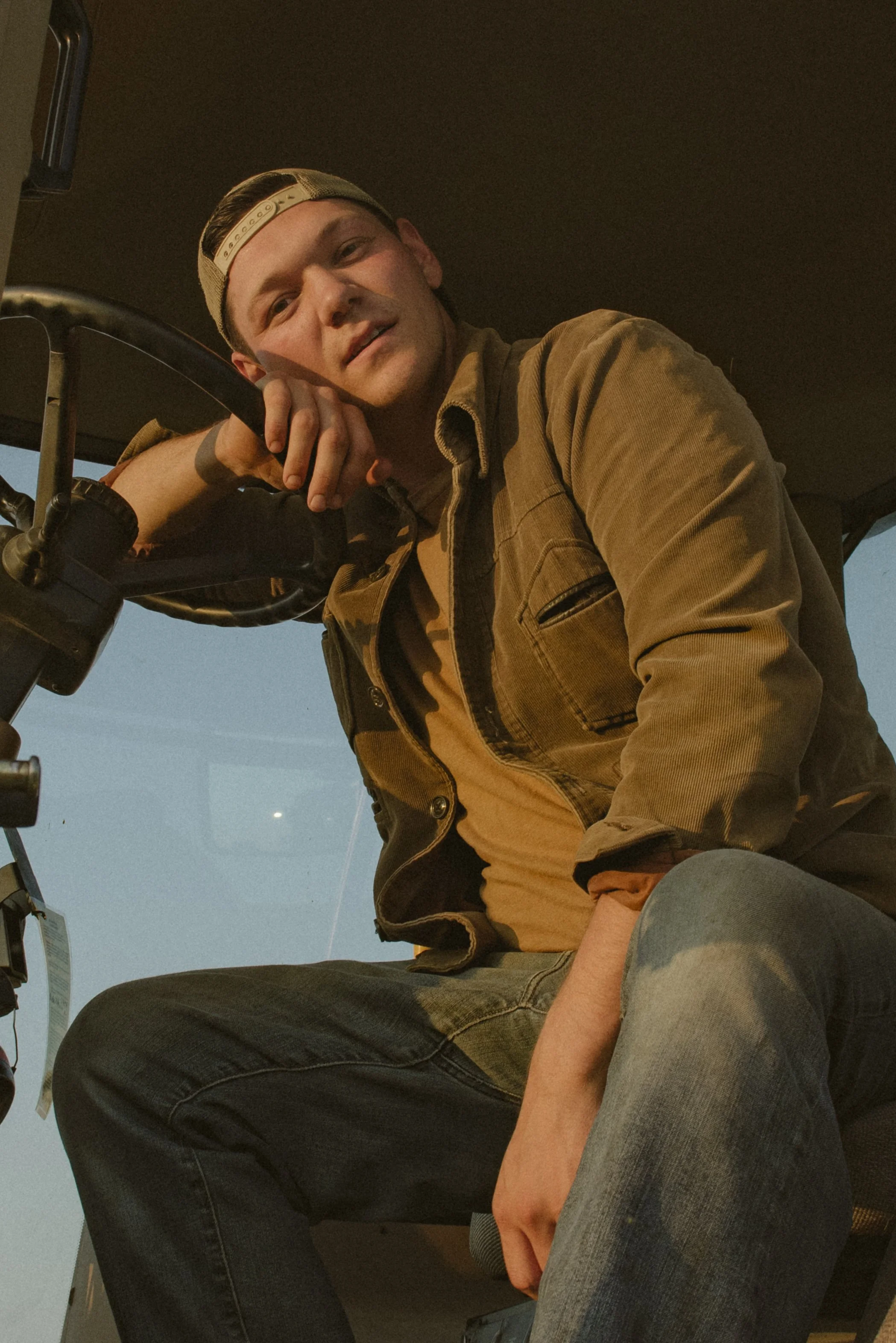 A young man sitting inside a vehicle, resting his arm on the steering wheel, wearing a tan shirt, brown jacket, a backward baseball cap, and jeans, looking at the camera with a relaxed expression.
