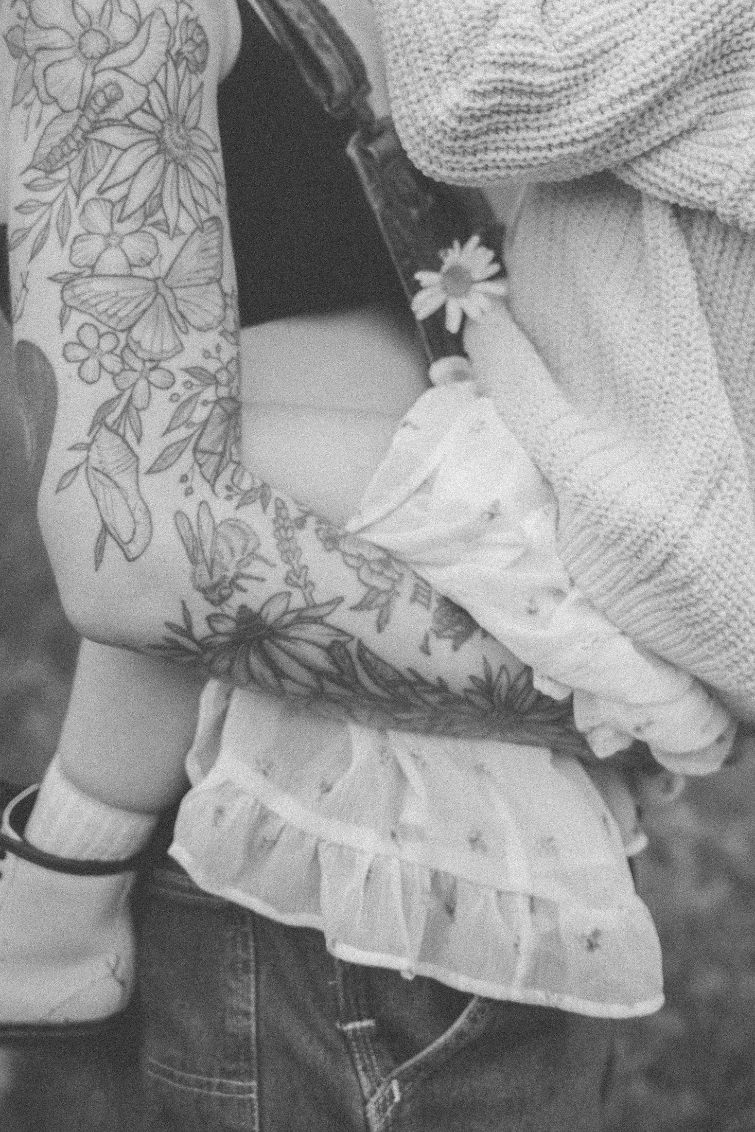 Close-up of a person's tattooed arm holding a baby swaddled in a blanket, with clothing featuring floral and butterfly patterns.