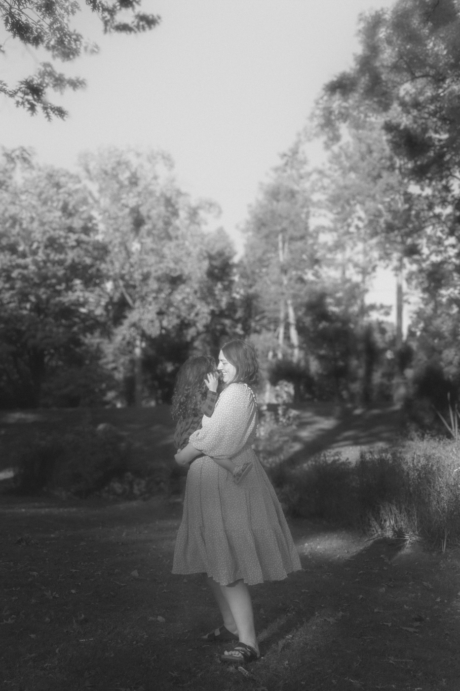 A woman holding a child and smiling outdoors in a park with trees and bushes
