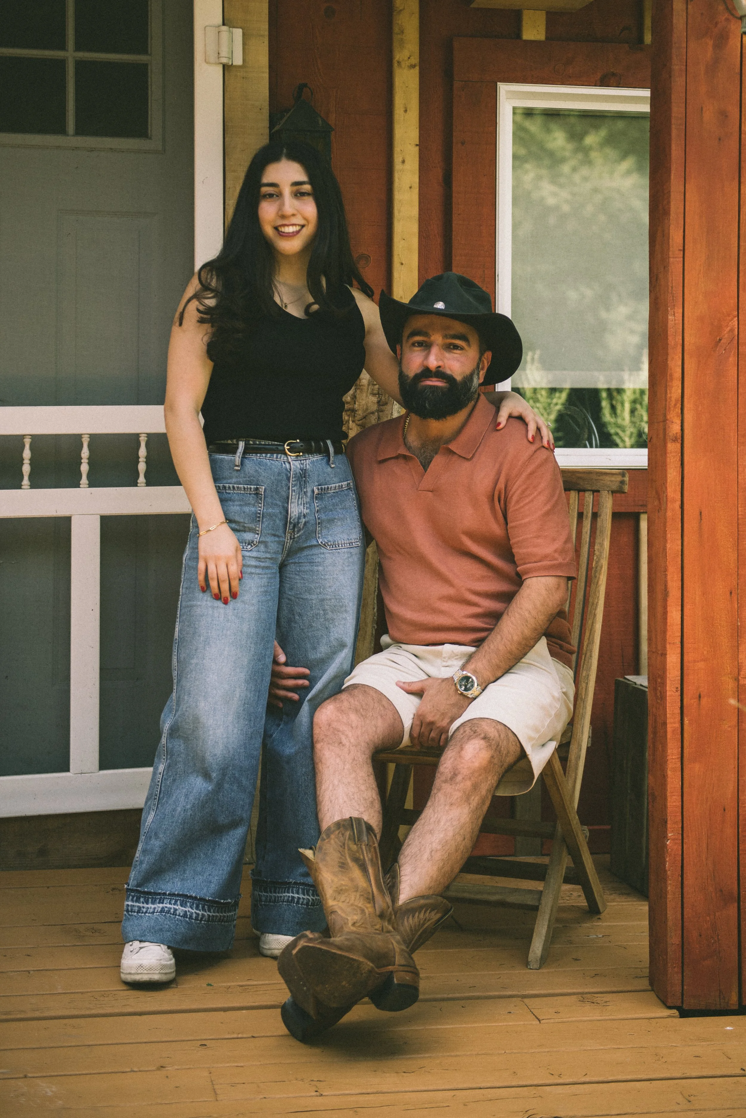 A young woman standing next to a seated man on a wooden porch, both smiling at the camera. The man is wearing a cowboy hat, a brown polo shirt, beige shorts, and cowboy boots. The woman has long dark hair, is wearing a black top and high-waisted blue