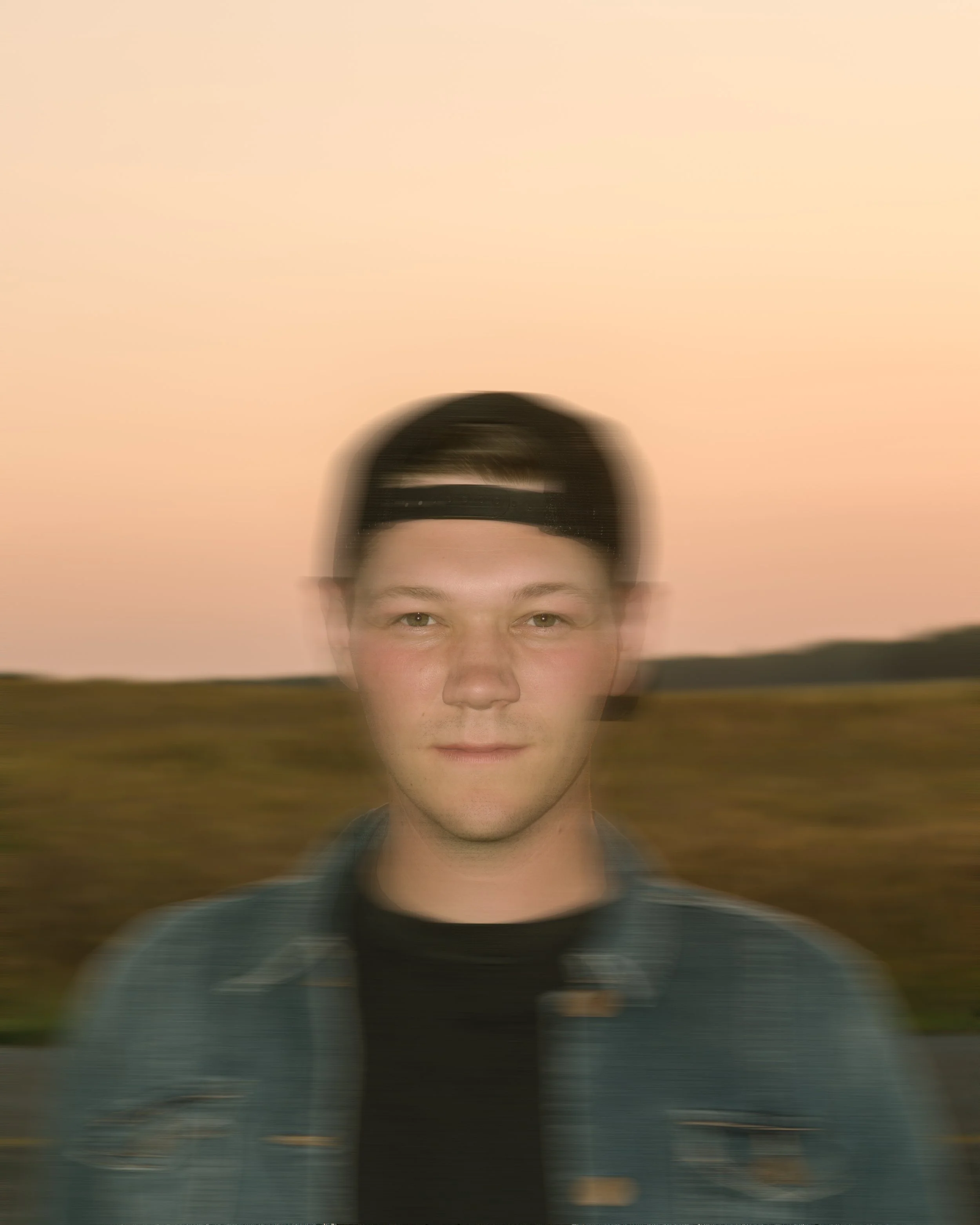 Young man wearing a backwards baseball cap, standing outdoors in a natural landscape during sunset, with a blurred background and slight motion blur on the face.