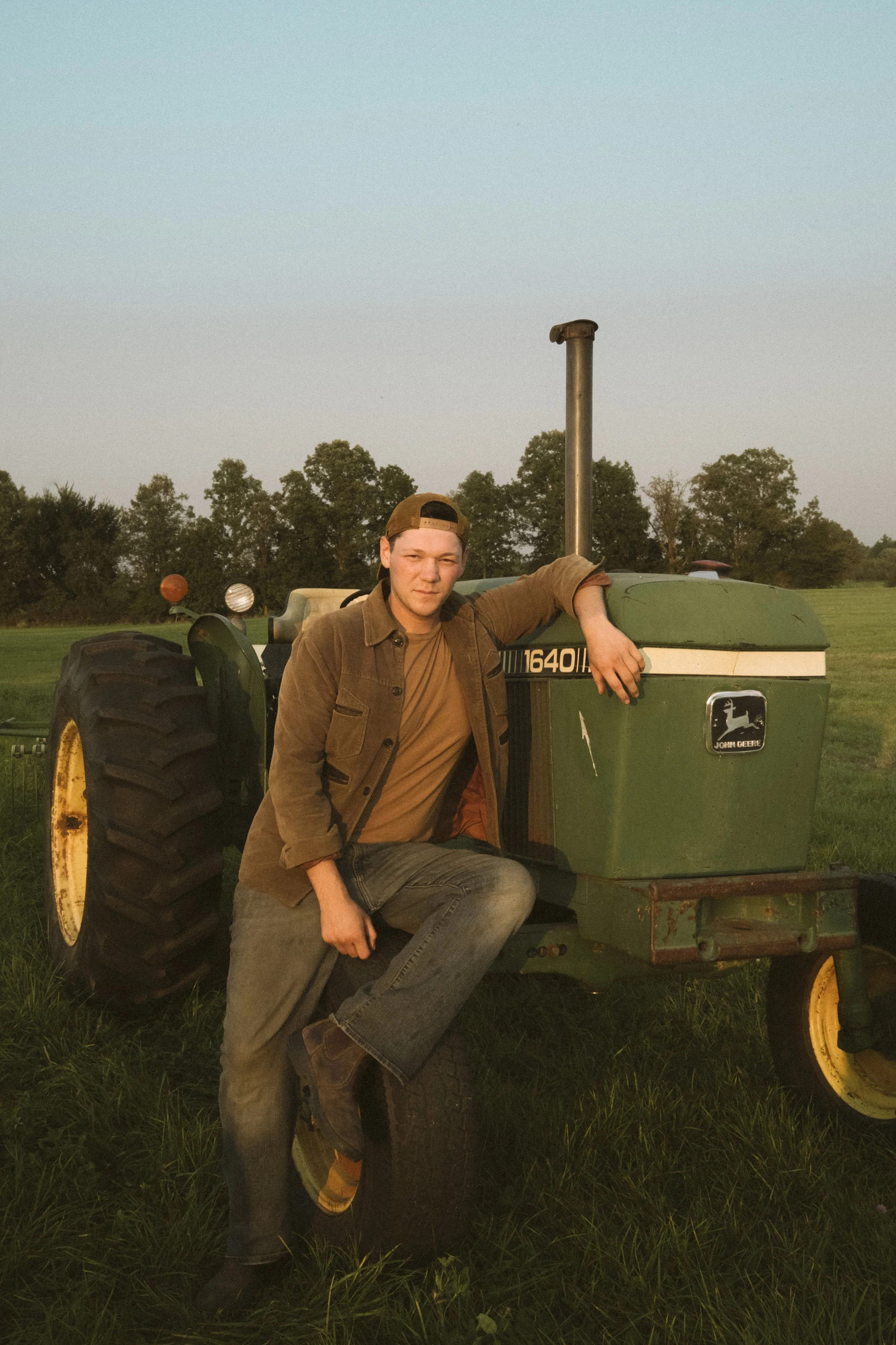 A young man in a brown jacket and jeans sitting on a rear tire of a green John Deere tractor in a grassy field during sunset.