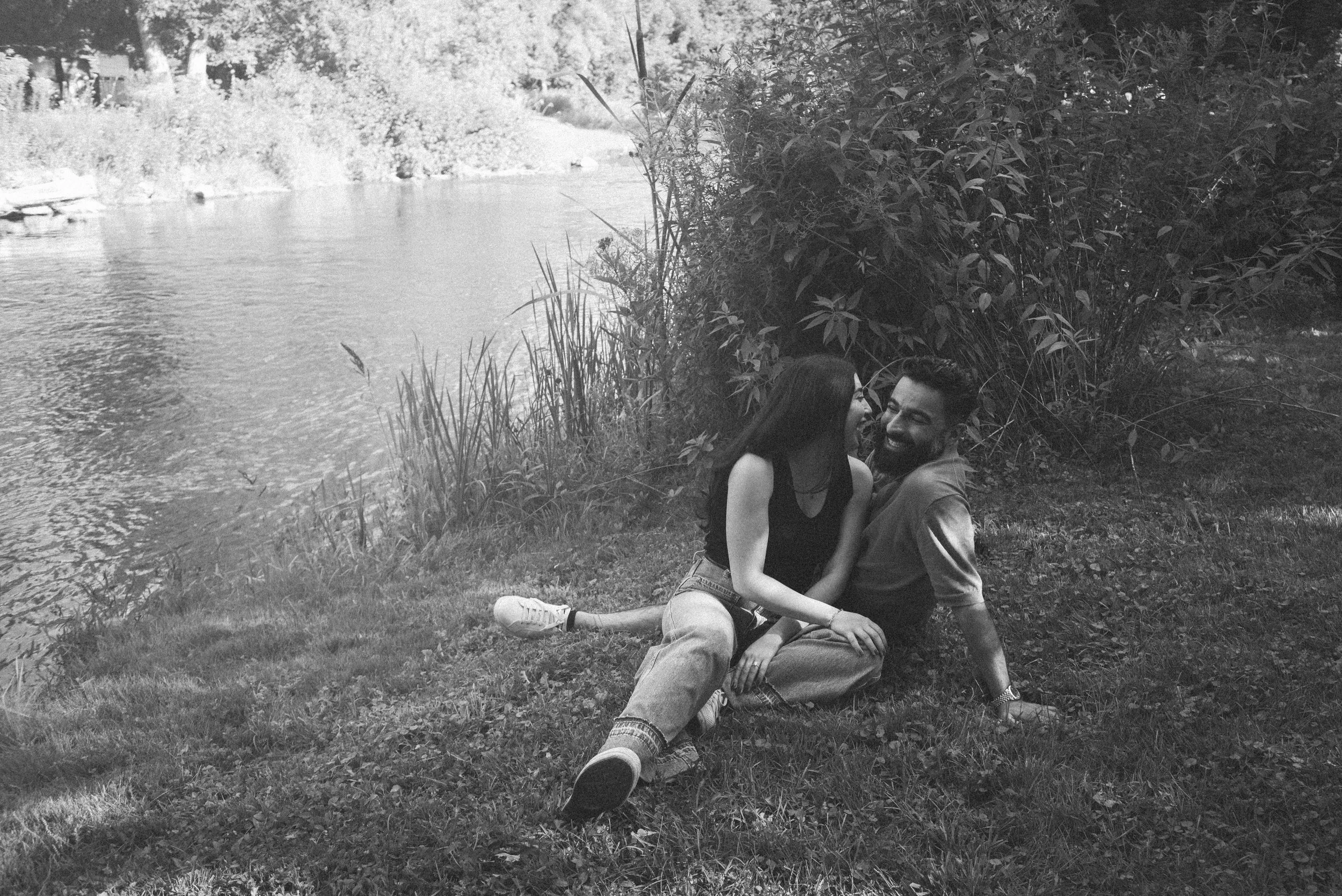 A couple sitting and smiling by a riverbank surrounded by greenery.