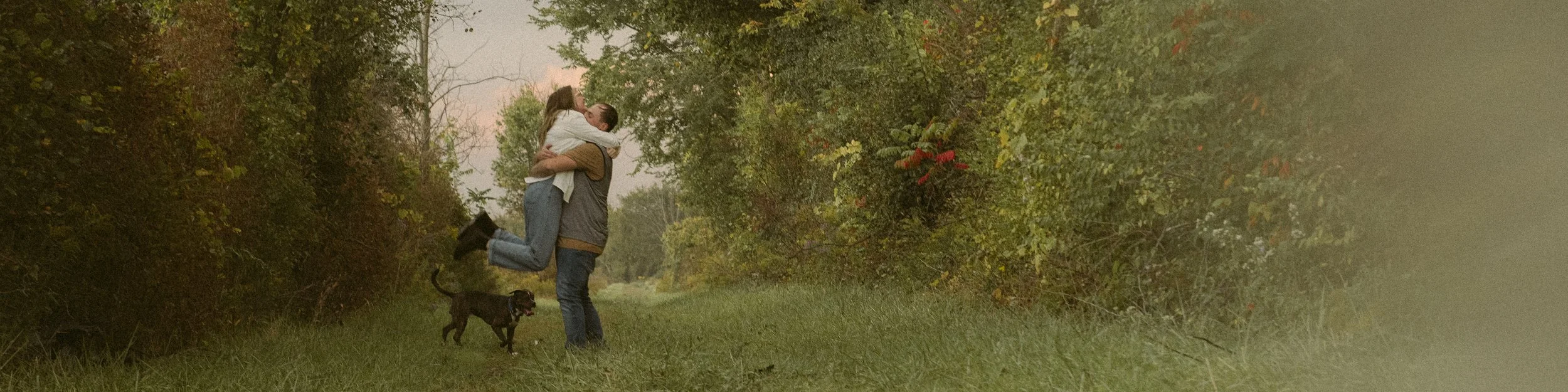 A couple is outdoors on a grassy path, with the man lifting the woman off the ground as they share a kiss. They are accompanied by a black dog, and there are trees with fall foliage surrounding them.