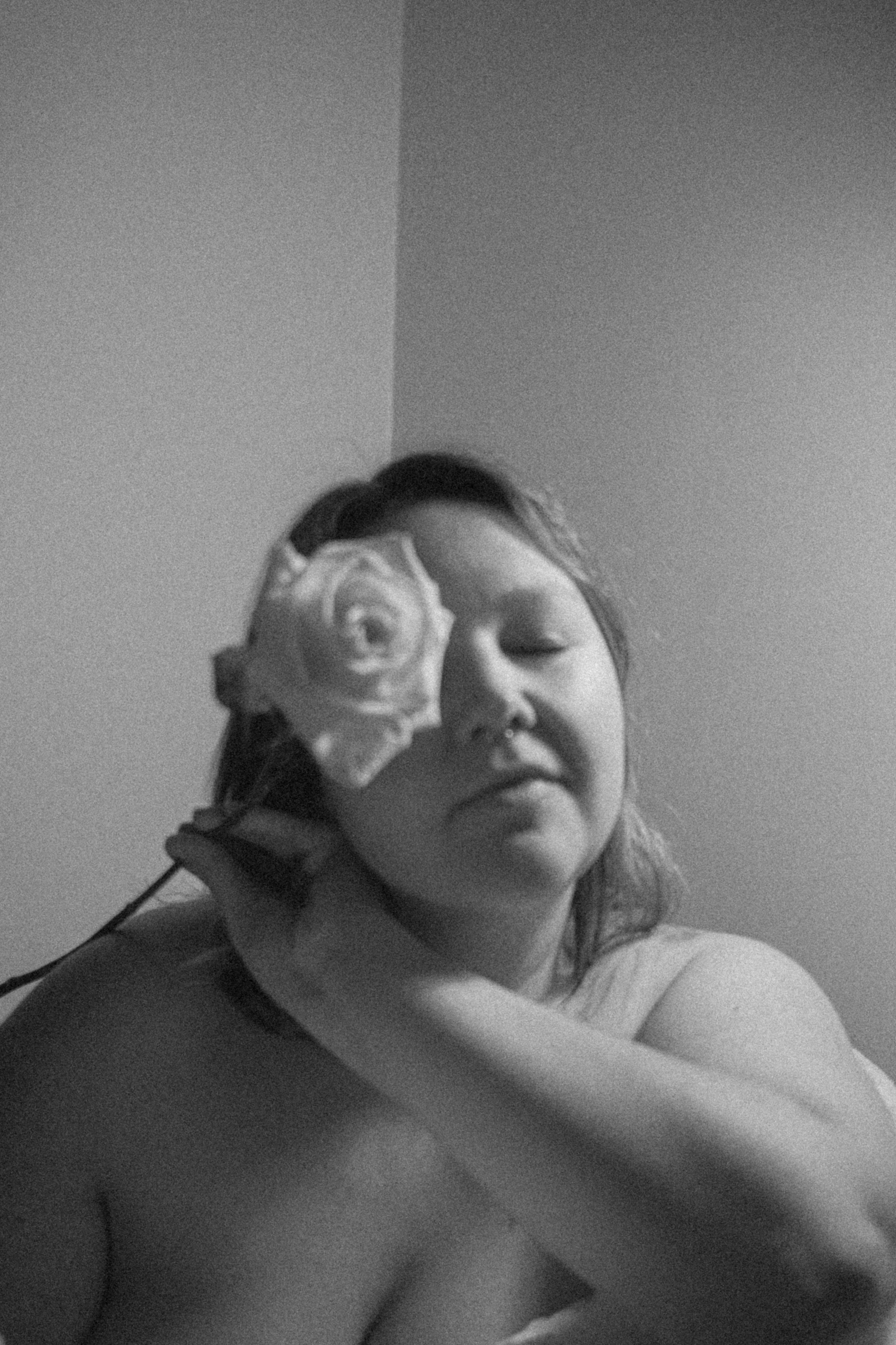 A person with medium-length wavy hair and a septum piercing holds a rose near their face, eyes closed in a relaxed pose, in a black and white photo.