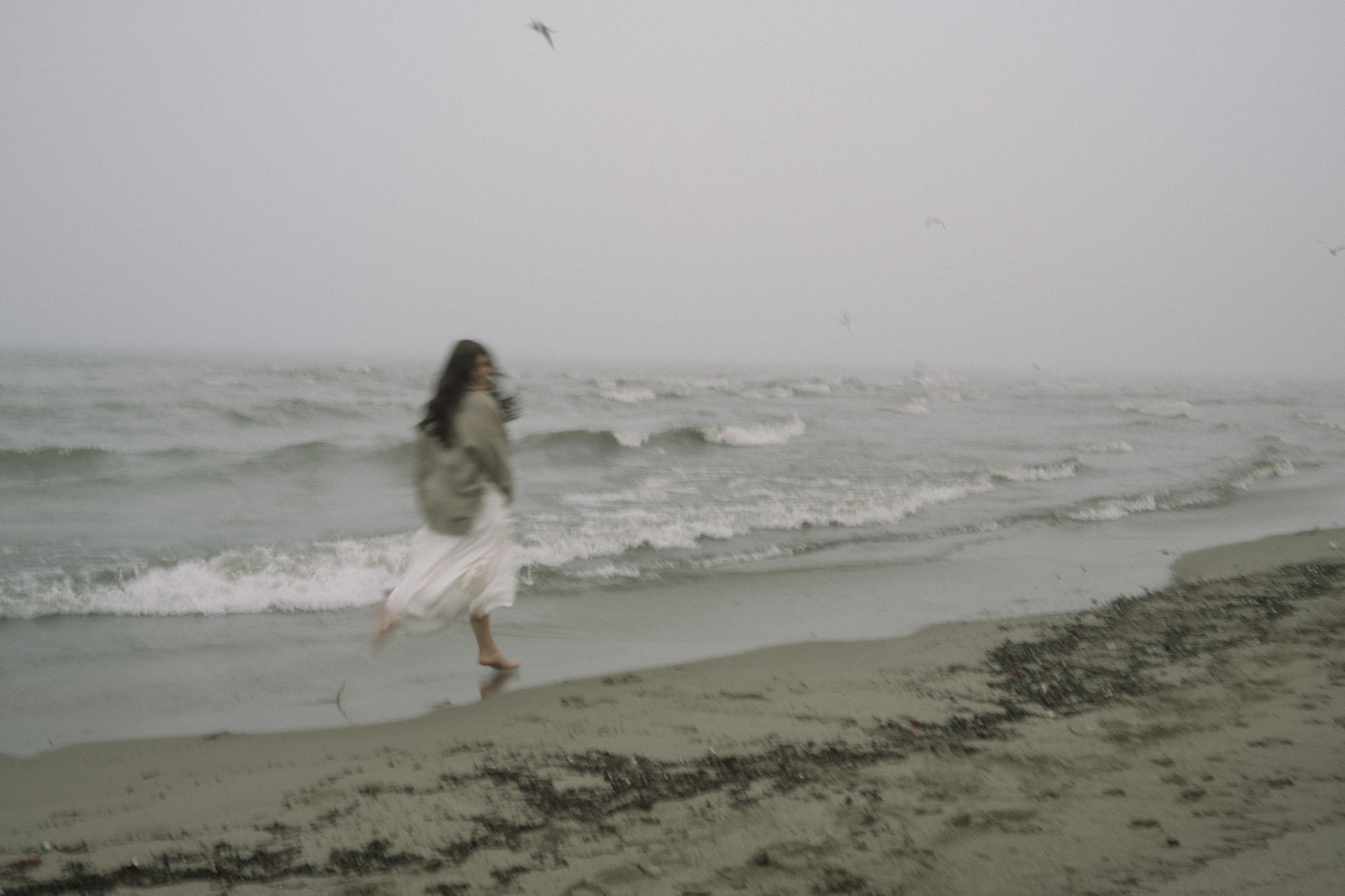 A woman with long dark hair walking along the shoreline on a foggy beach, wearing a beige jacket and a white skirt, with seagulls flying overhead.